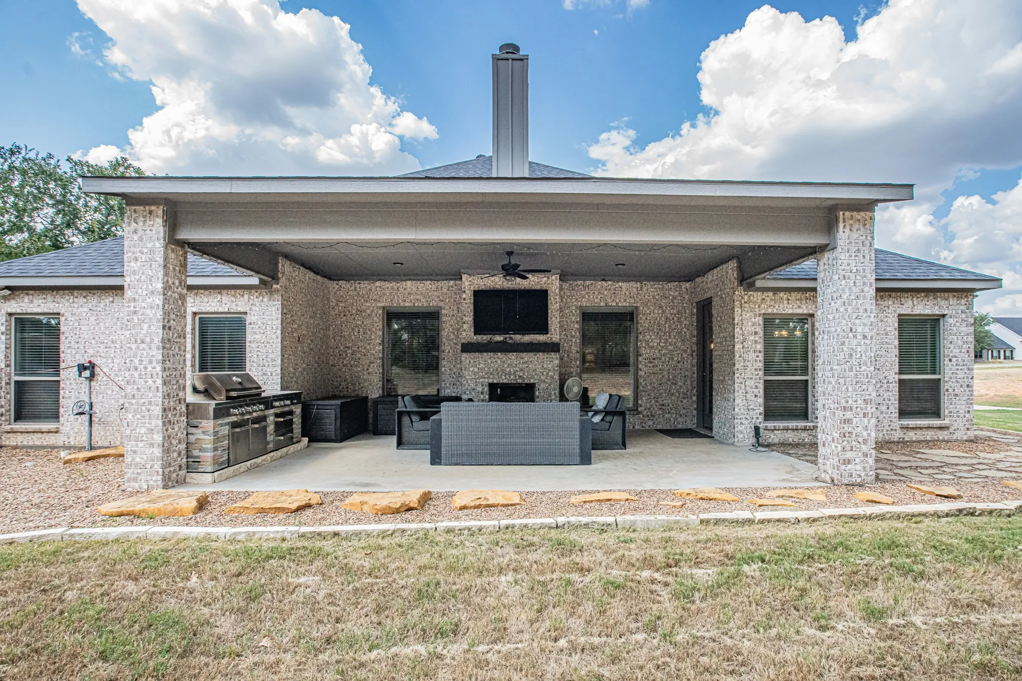 Expansive covered patio featuring a built-in outdoor kitchen, ceiling fan, and stone fireplace with a mounted television