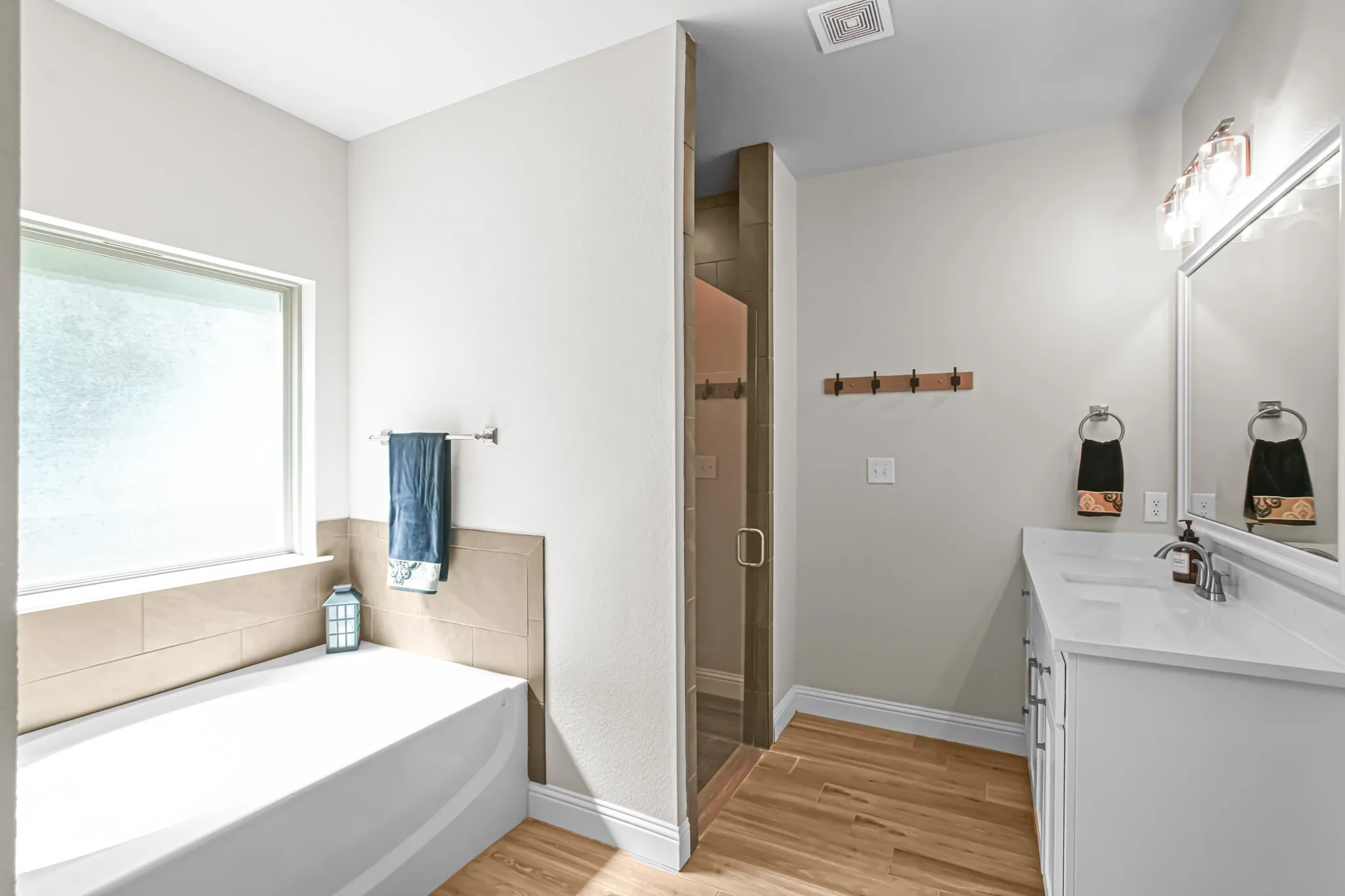 Bathroom featuring a built-in tub, tiled shower with glass enclosure, wood-finish flooring, dual vanity with white countertop, and a large wall-mounted mirror
