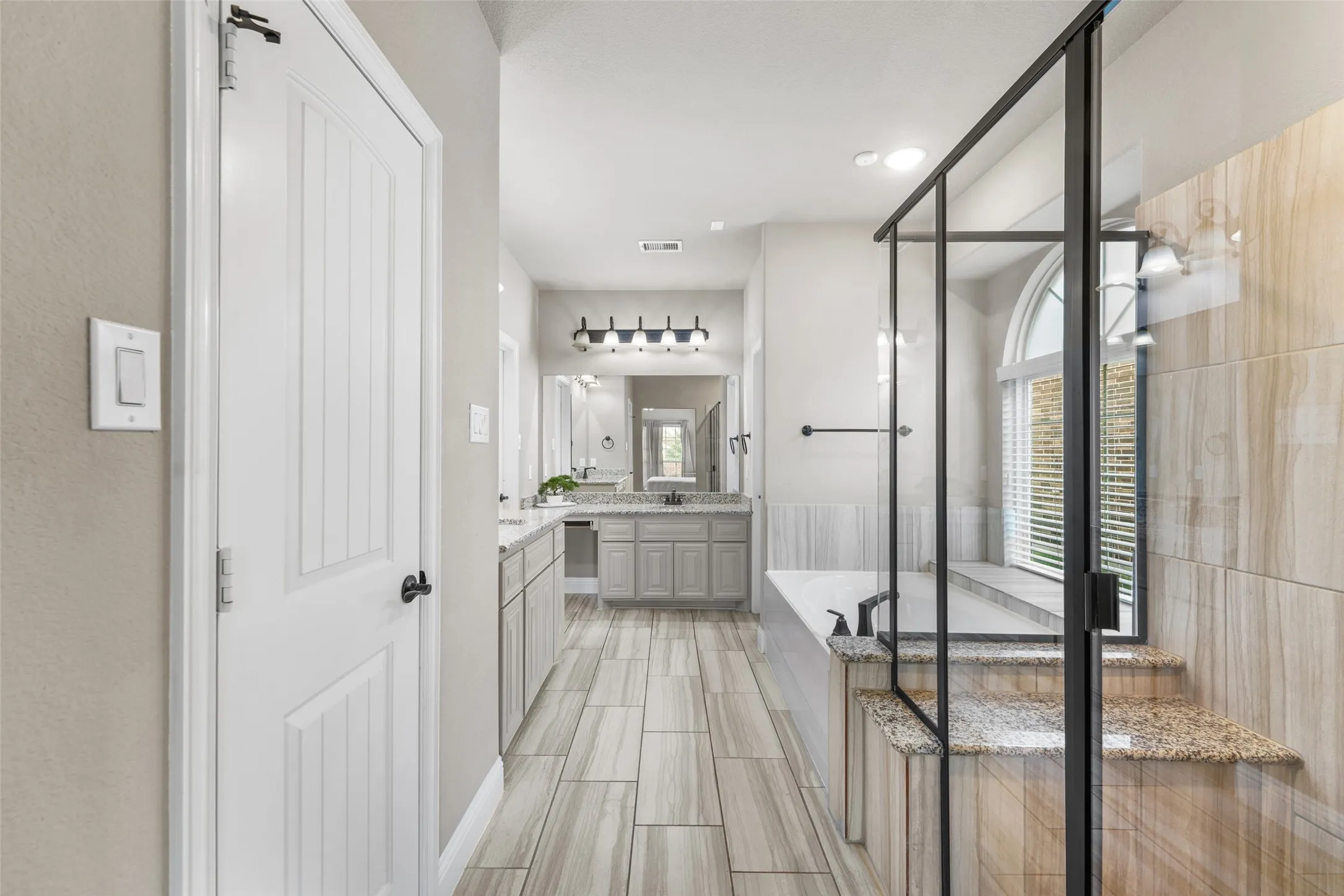 Porcelain tile flooring, dual vanities with granite countertops, separate soaking tub, and a frameless glass shower enclosure