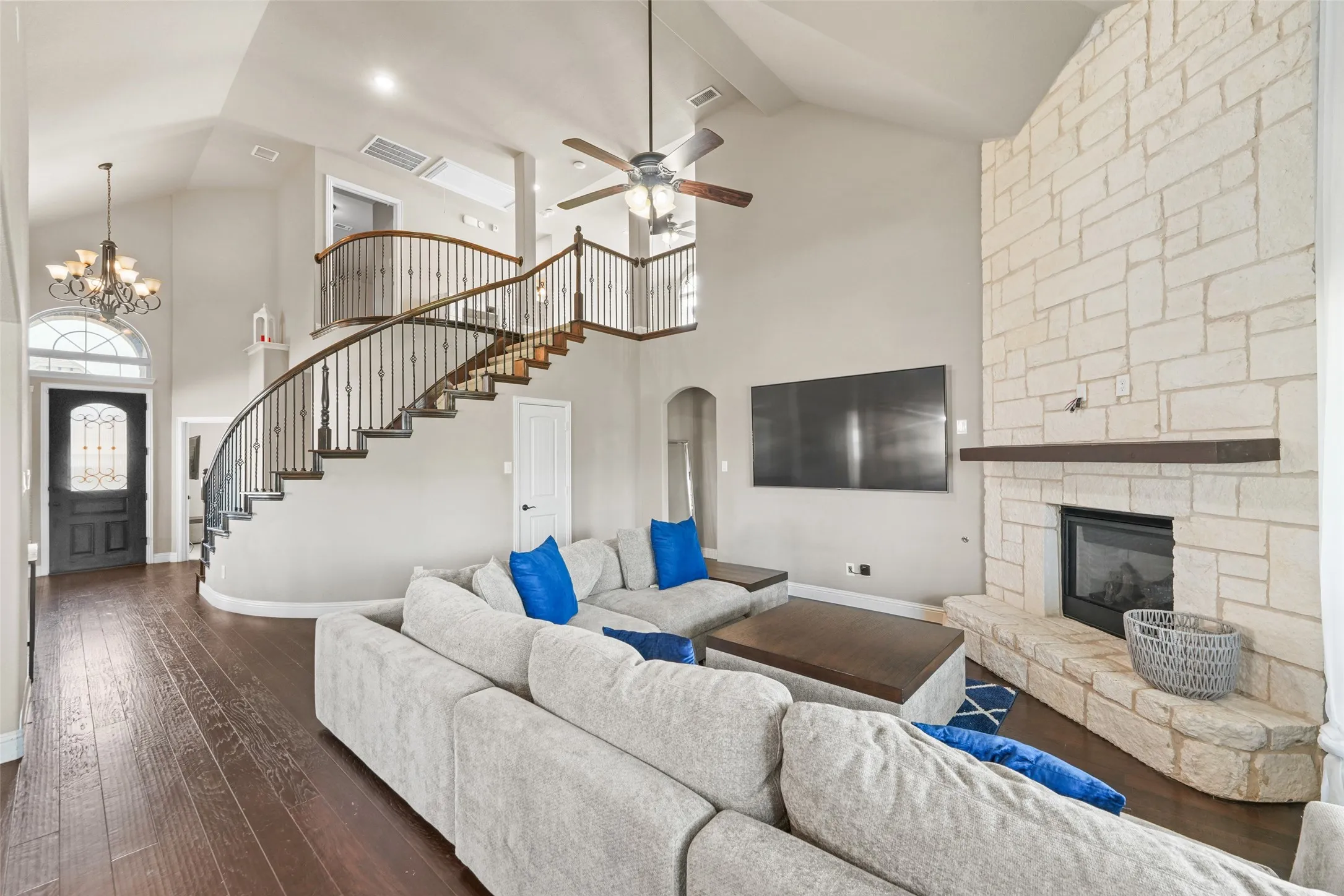 Vaulted ceiling with ceiling fan, wood-finish flooring, stone fireplace with a dark wood mantel, curved staircase with wood handrail and iron balusters, and a decorative front entry door with an arched transom window