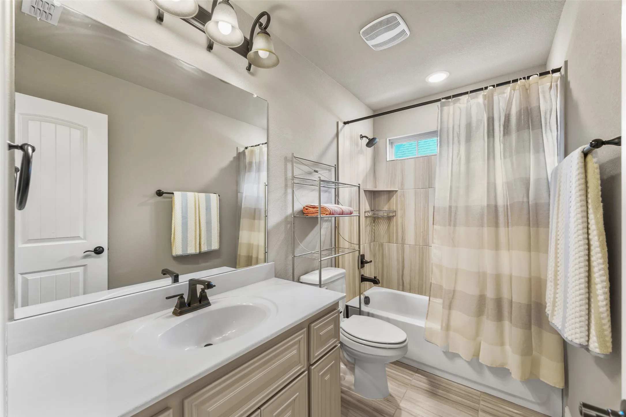 Single vanity with an integrated sink and bronze-finish faucet, full-width mirror, tile flooring, and a shower-tub combination with tile surround