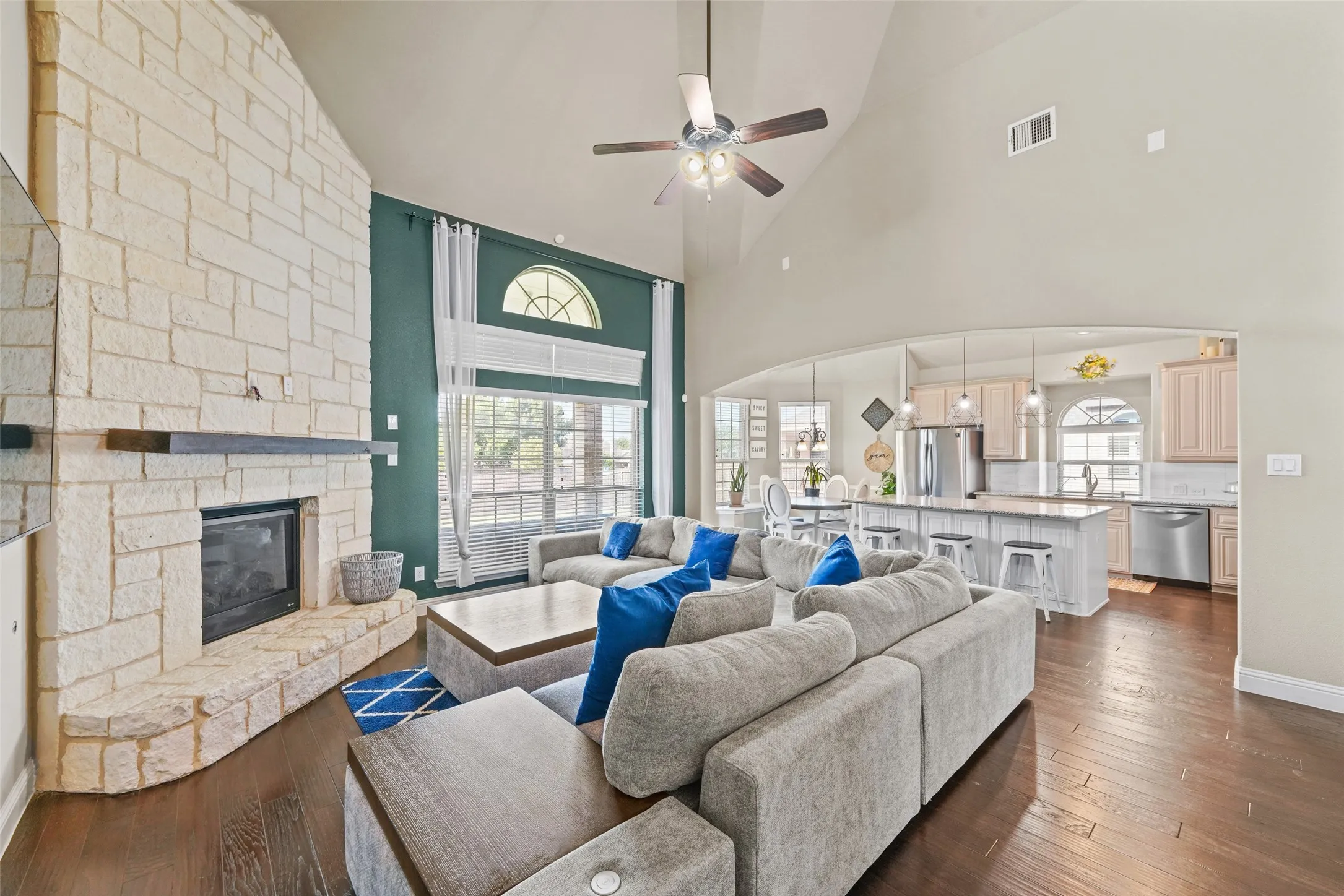Vaulted ceilings, a stone-clad fireplace with a dark mantel, wood-finish flooring, and a kitchen with stainless steel appliances