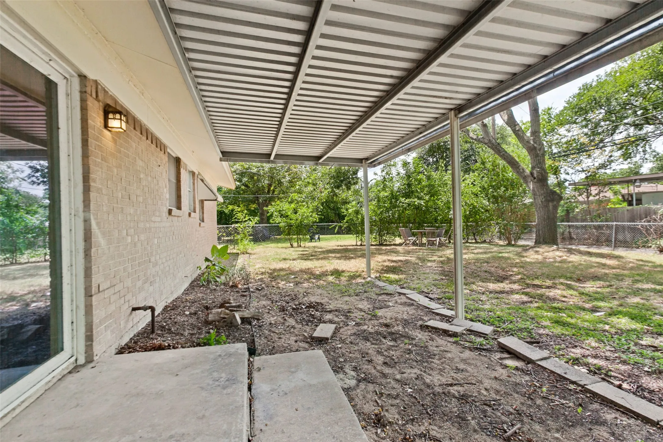 Covered patio featuring a metal roof structure, concrete patio area, brick exterior, and a spacious yard with mature trees