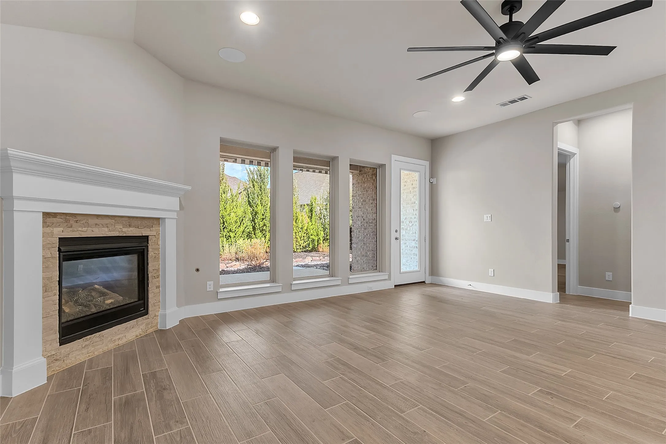Living room with a gas fireplace, hardwood flooring, and large windows overlooking a landscaped yard, illuminated by recessed lighting and ceiling fan.