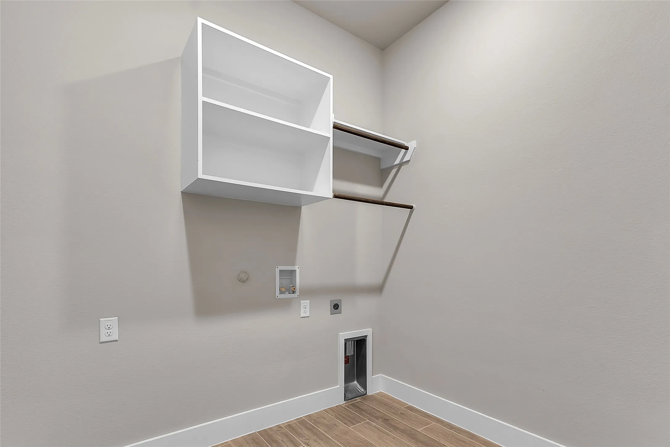 Laundry room with white shelving, wooden rods, light-colored walls, and a utility hookup on the floor for appliances.