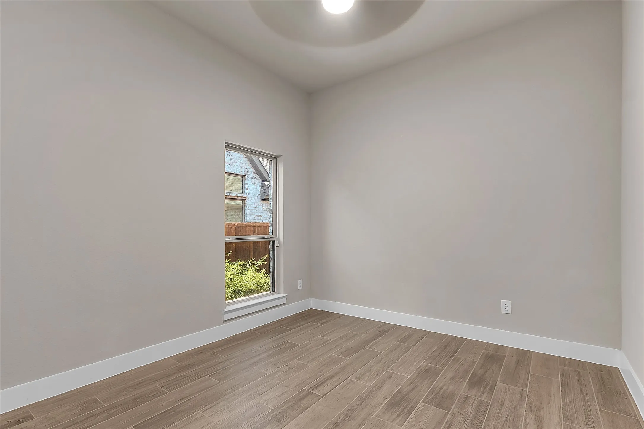 Empty room with light gray walls, a window allowing natural light, and wood-look tile flooring.