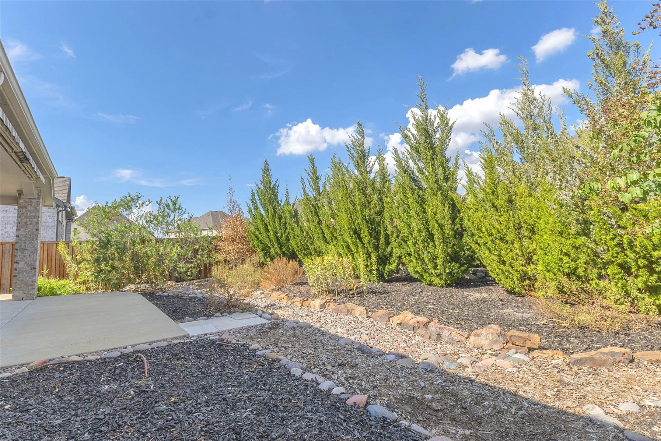 Backyard landscape with natural stone borders, landscaped with evergreen trees, gravel beds, and a concrete patio area for outdoor relaxation.