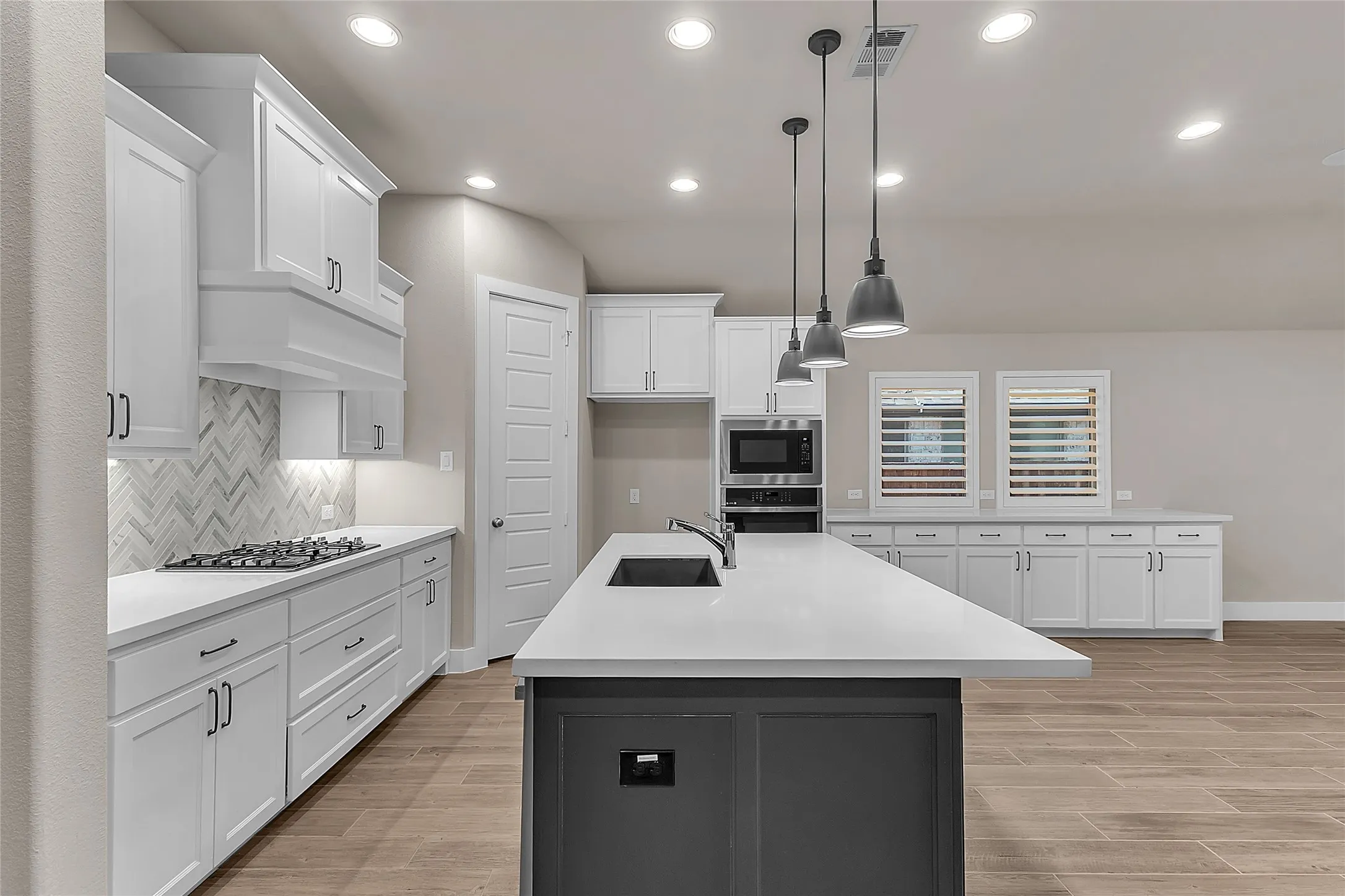 Open-plan kitchen with white cabinetry, quartz countertops, stainless steel appliances, herringbone tile backsplash, and recessed lighting above an island.