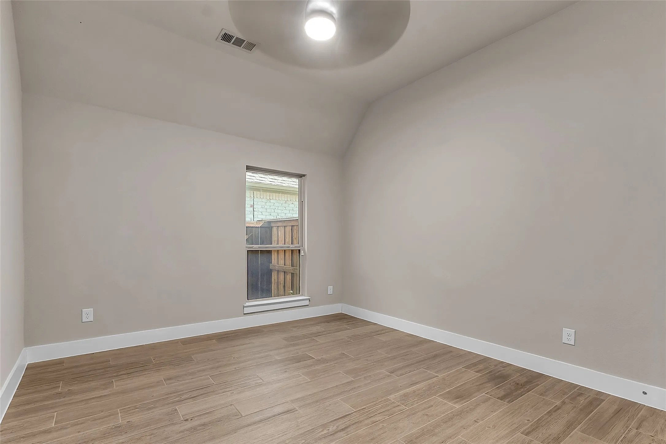 Empty room with neutral walls, wood-look tile flooring, and a window providing natural light with a vaulted ceiling