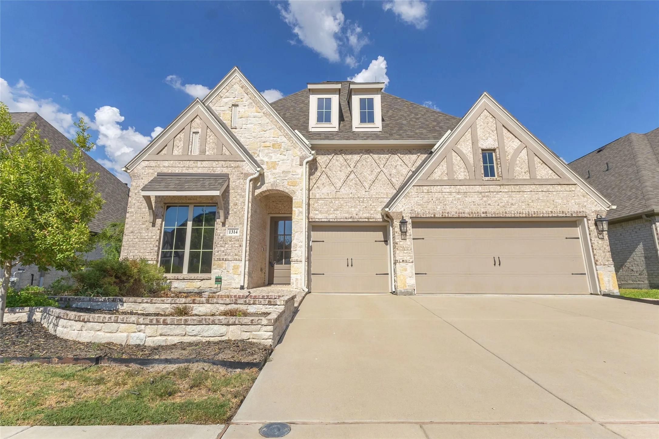 Three-car garage with light beige stone exterior, bronze hardware, and symmetrical layout, complemented by a paved driveway and landscaping.
