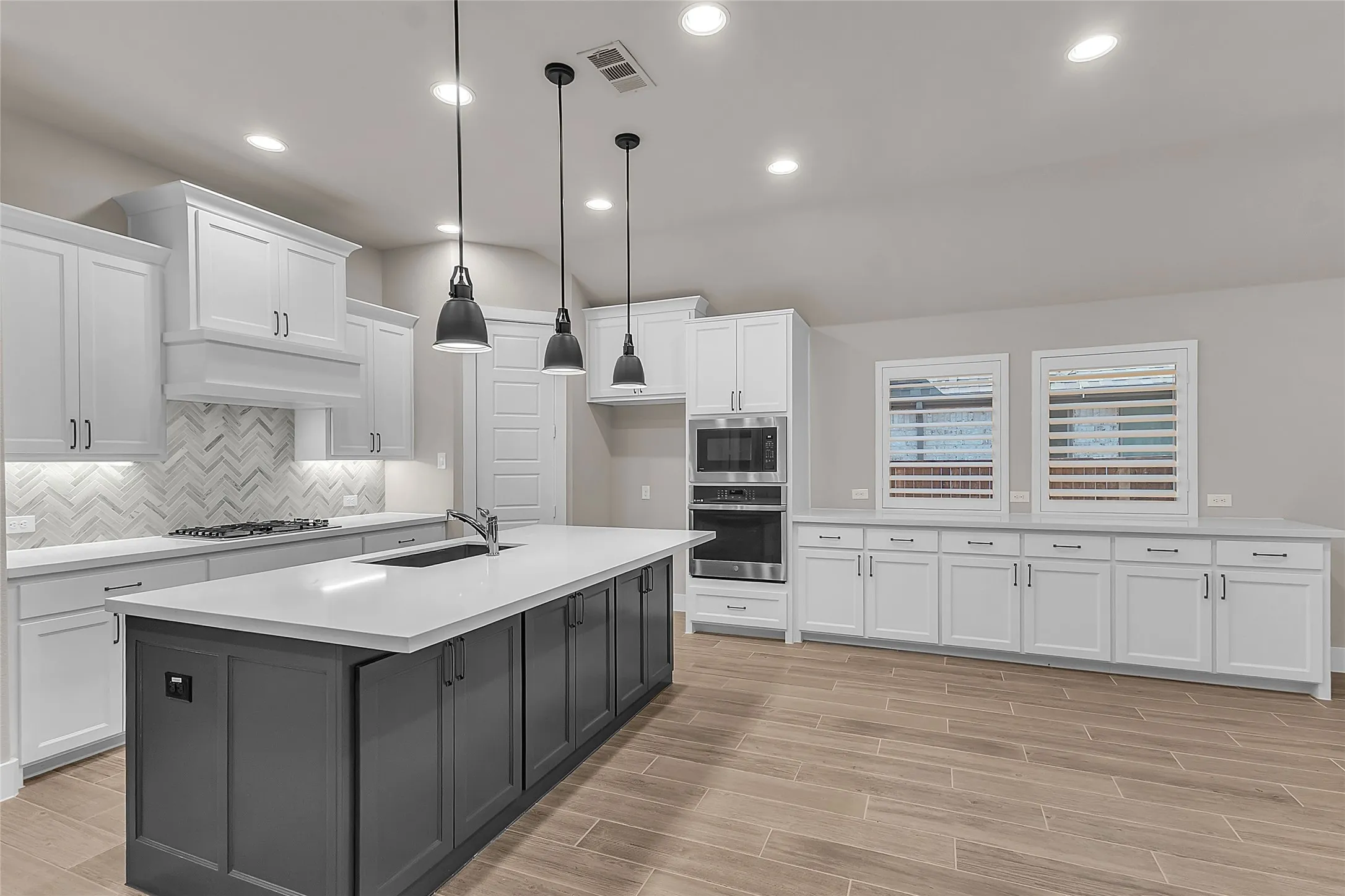 Open-plan kitchen with gray and white cabinetry, quartz countertops, stainless steel appliances, pendant lighting, and herringbone tile backsplash