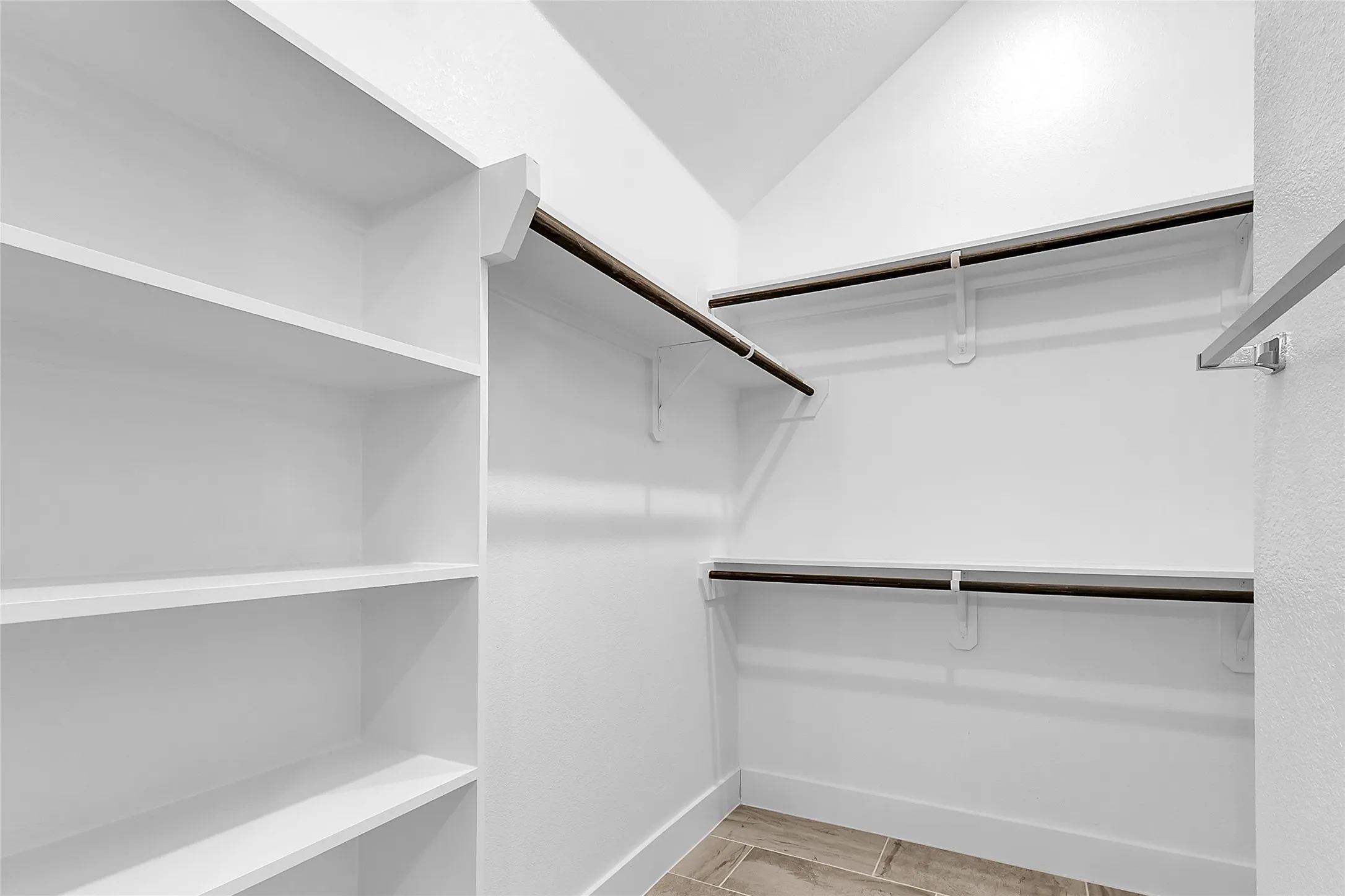 Walk-in closet with white walls, wooden hanging rods, and tile flooring, highlighted by a vaulted ceiling and ample storage space.