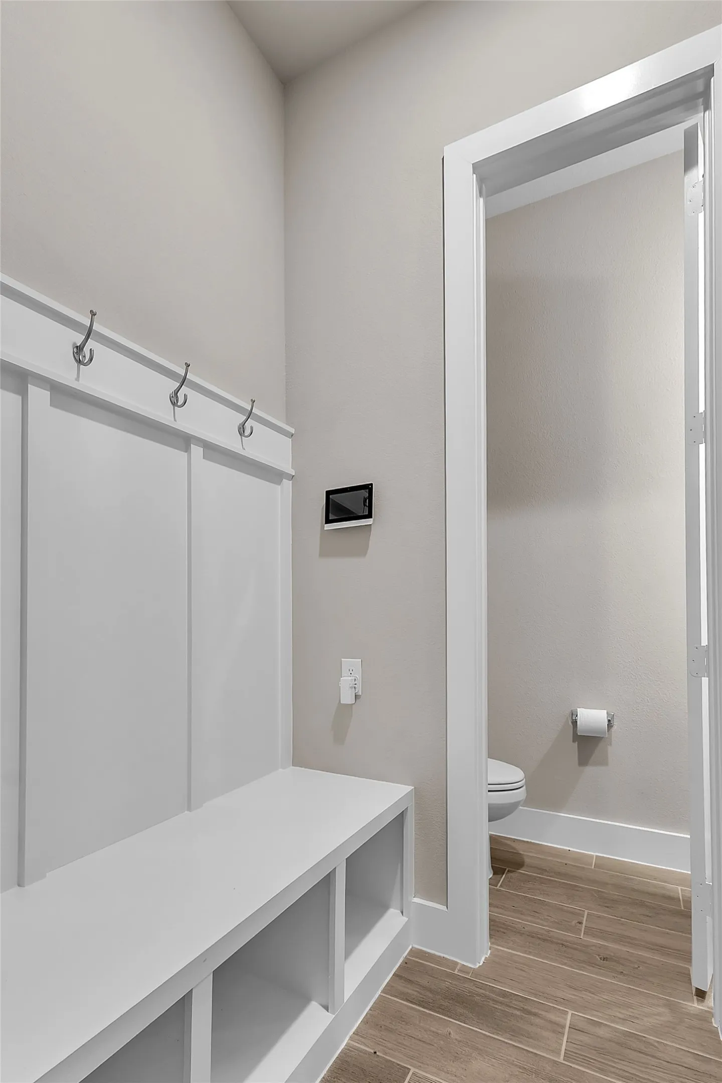Mudroom with built-in bench and storage cubbies, white shiplap walls, and light wood plank flooring adjacent to a powder room with a toilet and wall-mounted paper holder
