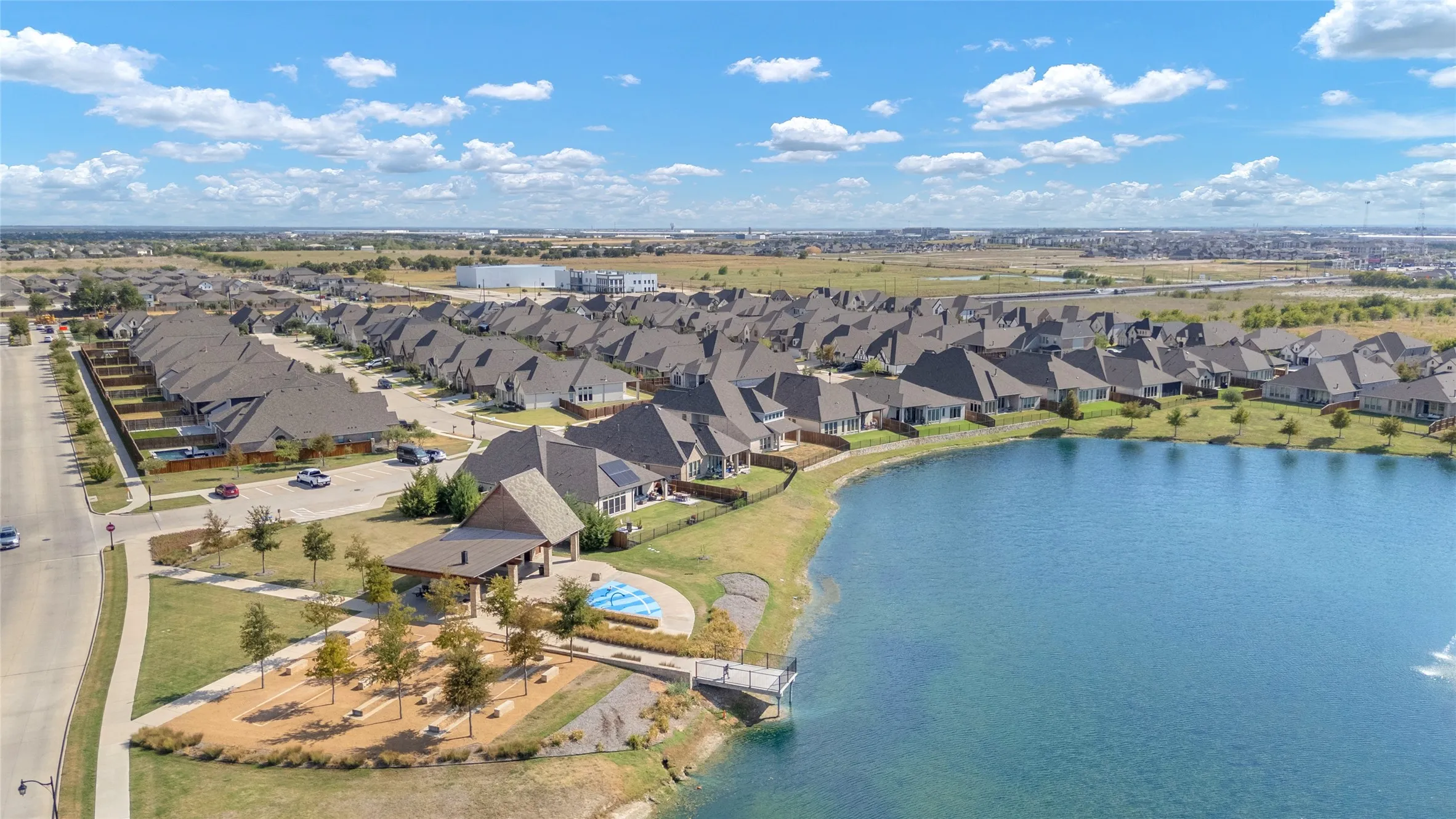 Aerial view of residential community with lake, landscaped yards, and a mix of single-family homes with asphalt roofs and green spaces
