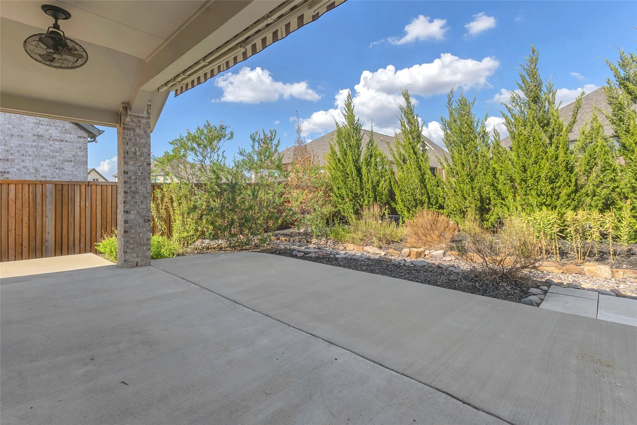Covered back patio with concrete flooring, bordered by landscaped gravel and mature shrubs, providing a private outdoor space for relaxation.