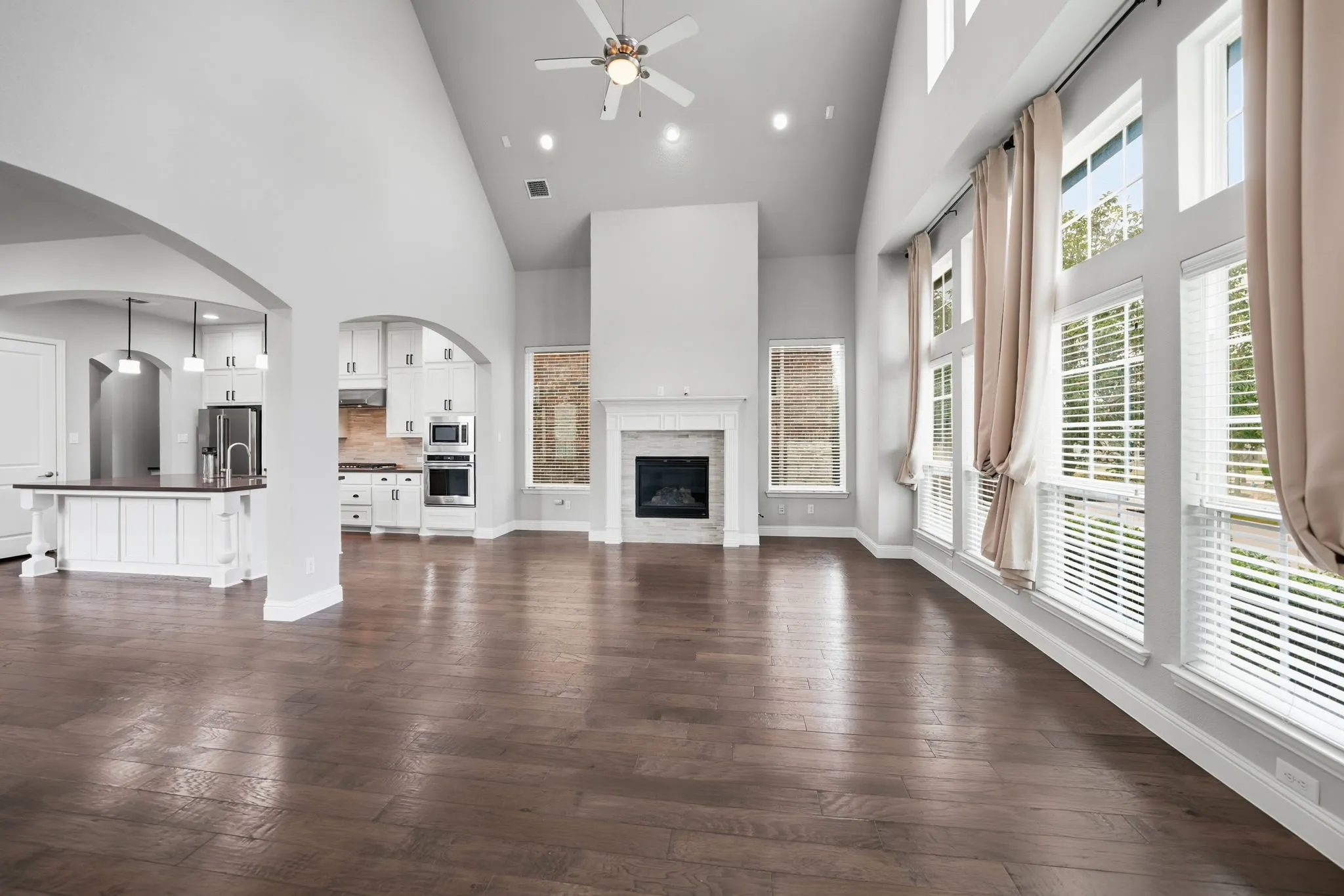 Vaulted ceiling with recessed lighting, wood-finish flooring, two-story windows with blinds, built-in fireplace with white mantel, kitchen island with pendant lighting