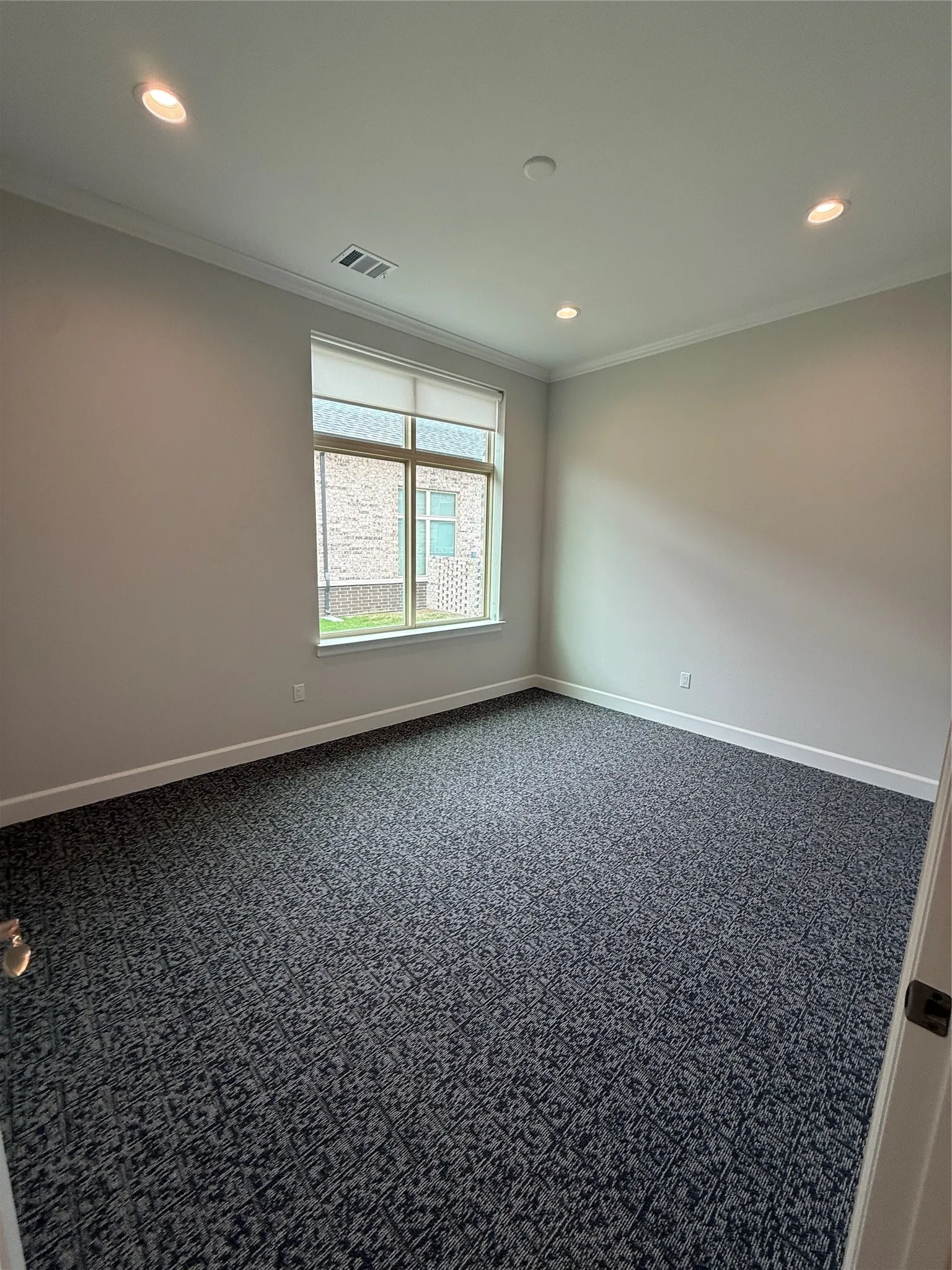 Recessed ceiling lighting, patterned carpet flooring, a large window with a roller shade, and white crown molding