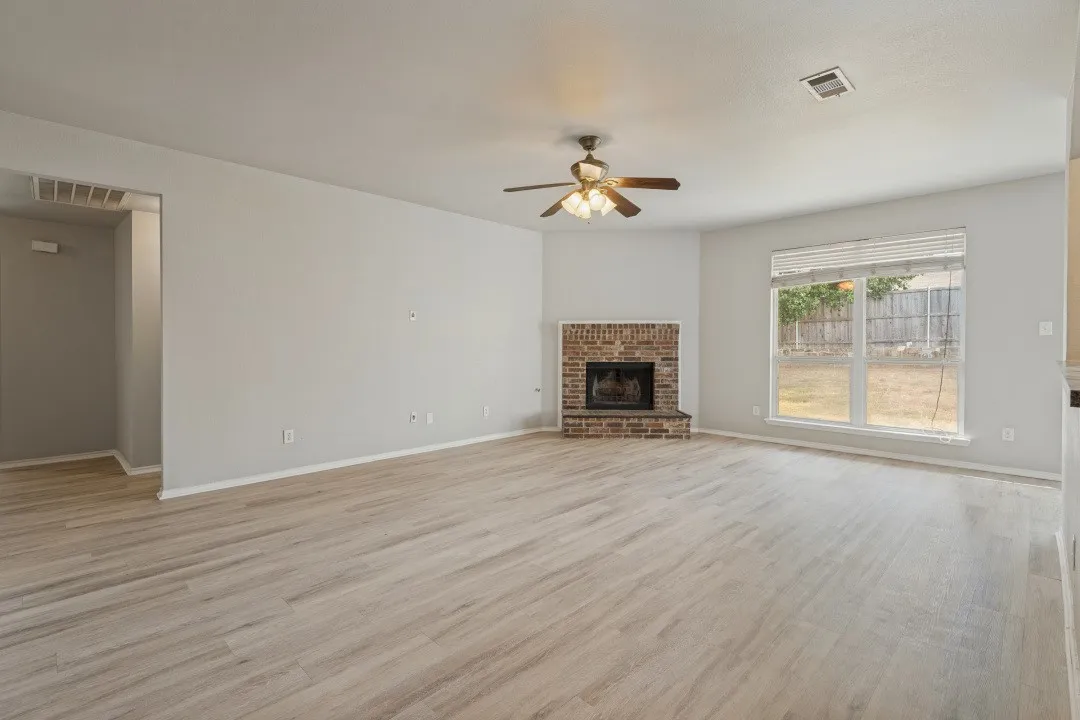 Spacious room featuring wood-finish flooring, a brick fireplace with a hearth, a ceiling fan with integrated lighting, and a large window overlooking the backyard