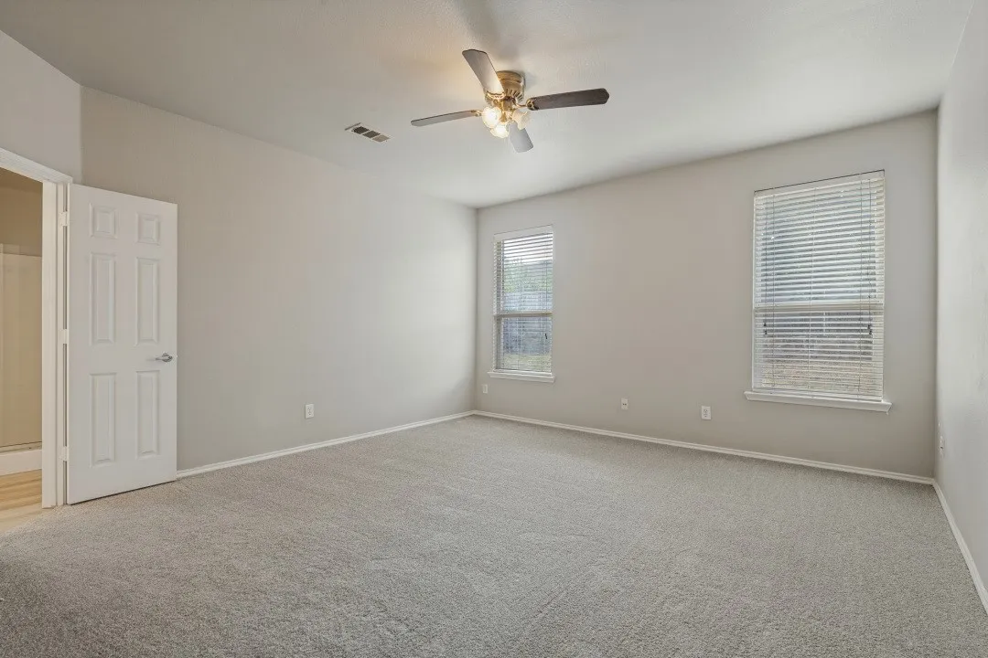 Spacious room featuring neutral carpeting, two windows with blinds, a ceiling fan with integrated lighting, and a paneled door