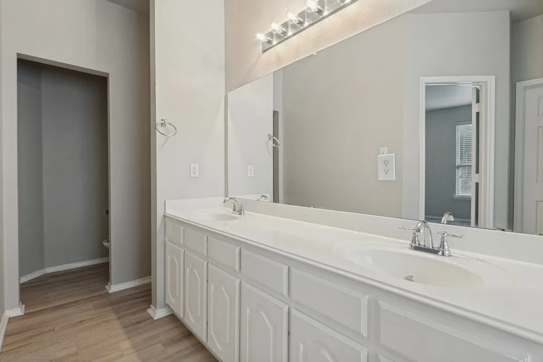 Dual vanity with integrated sinks, white cabinetry, wood-finish flooring, and an expansive mirror with overhead lighting