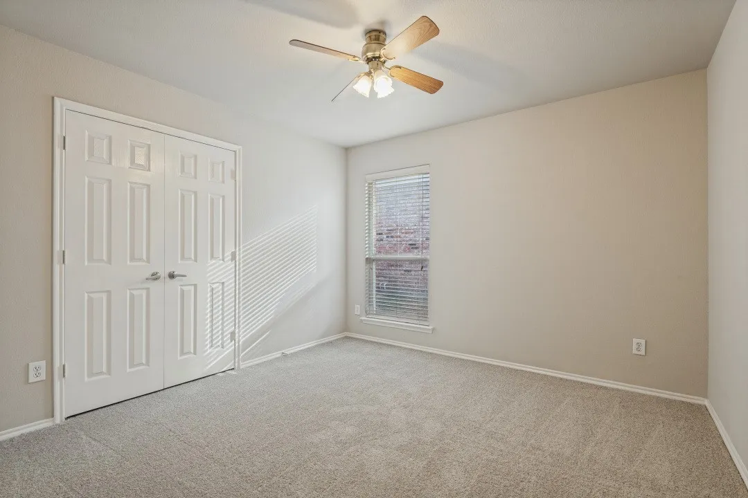 Neutral-toned carpeting, ceiling fan with light fixture, window with blinds, and paneled closet doors