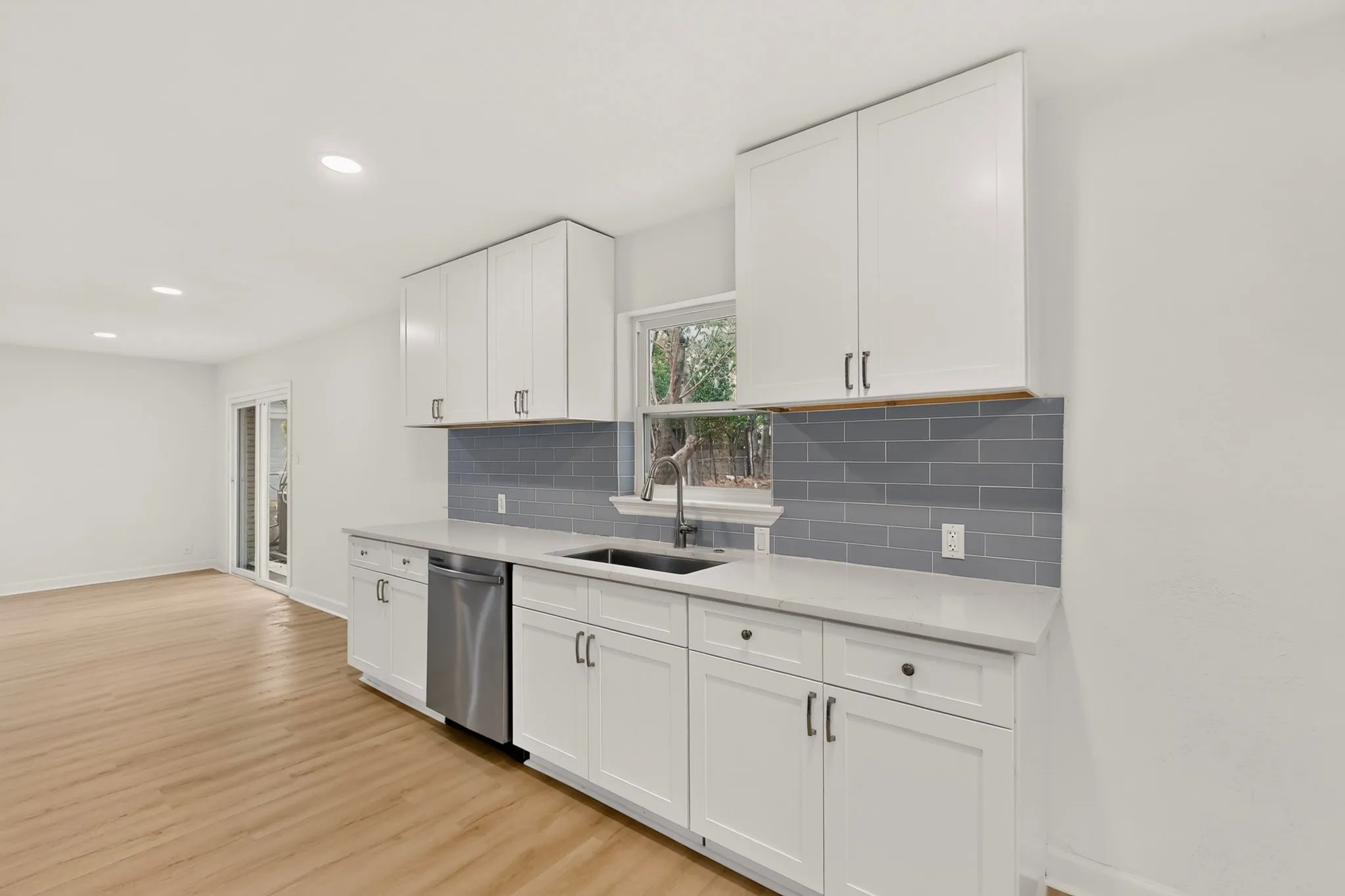 Modern kitchen featuring white shaker cabinetry, stainless steel dishwasher, light-toned wood-finish flooring, gray subway tile backsplash, and light-toned countertops