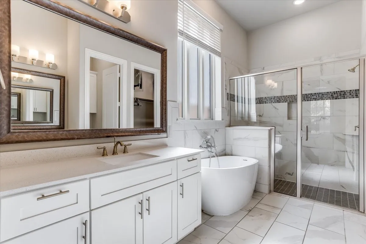 Dual-sink vanity with white cabinetry and light-toned countertop, freestanding soaking tub, glass-enclosed shower with tiled walls, and large framed wall mirror
