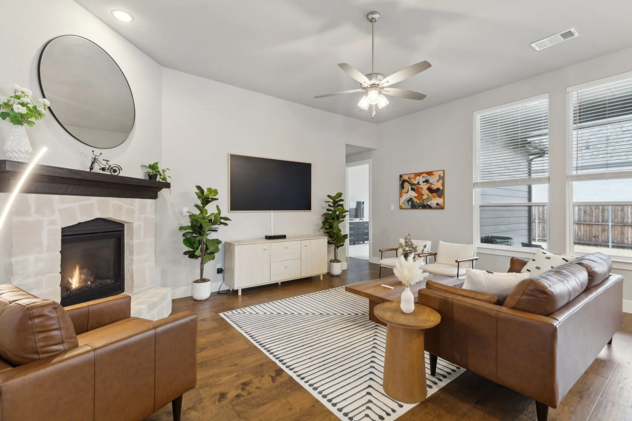Stone-clad fireplace with dark wood mantle, high ceilings, wood-finish flooring, ceiling fan, and large windows
