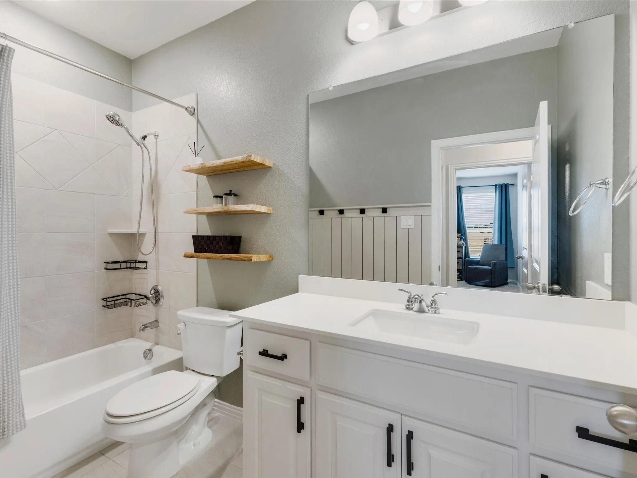 White vanity with an integrated sink and chrome faucet, white tub with a shower head and handheld sprayer, wood-finish floating shelves, and wainscoting detail