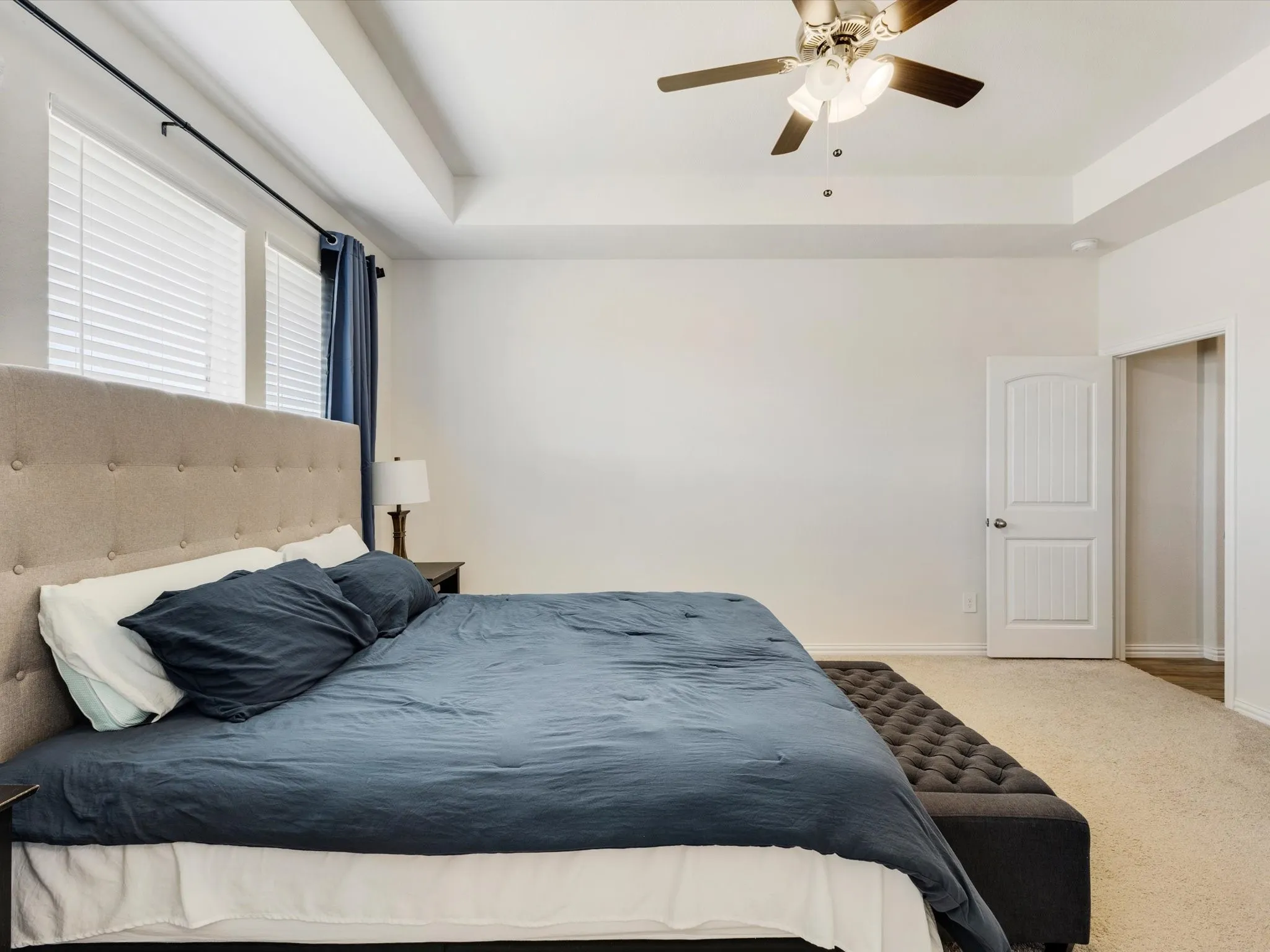 Tray ceiling with a ceiling fan light fixture, two windows with blinds, and white paneled interior doors