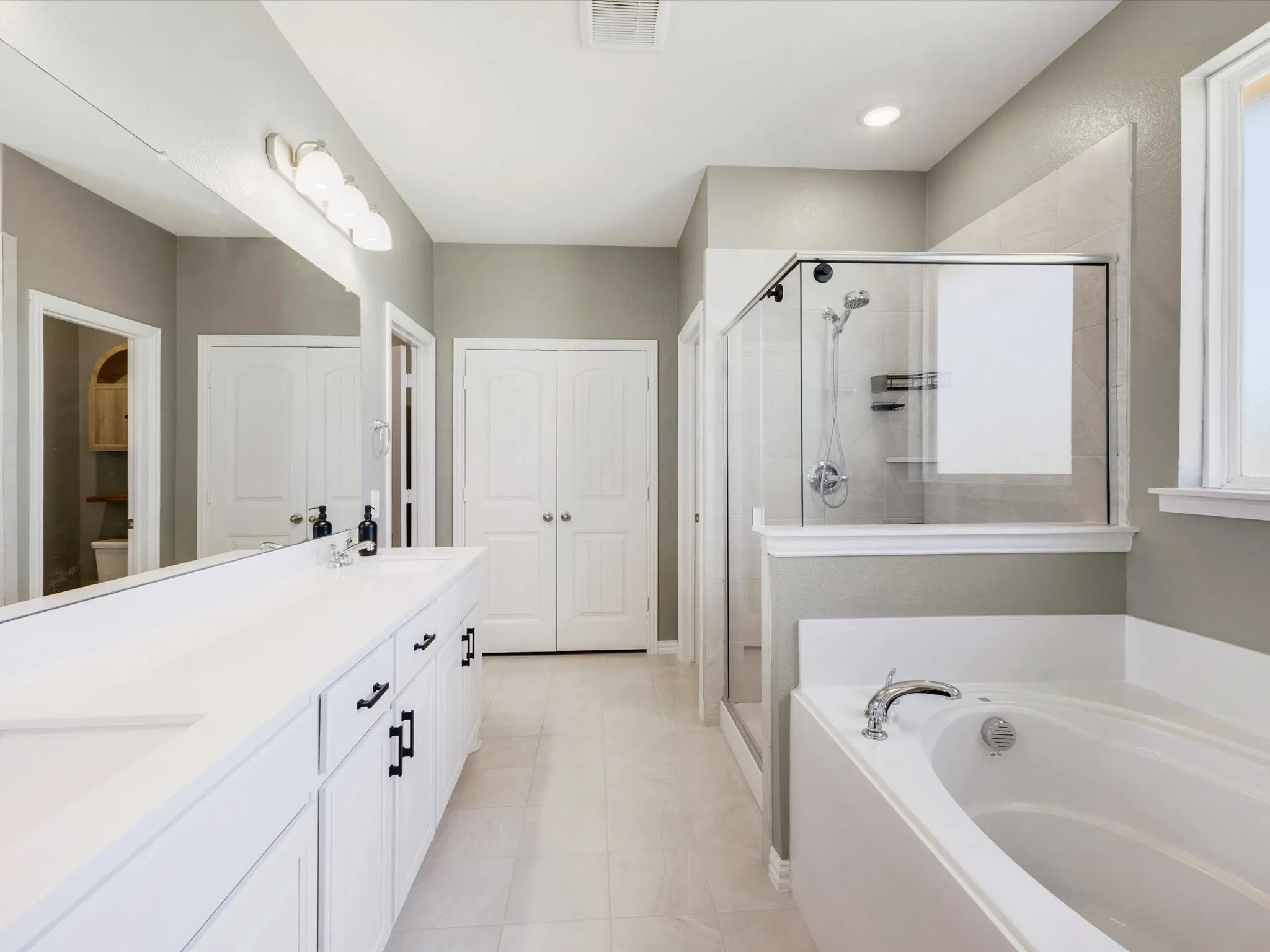 Dual vanity with white cabinetry and black hardware, glass-enclosed shower, built-in soaking tub, and tile flooring