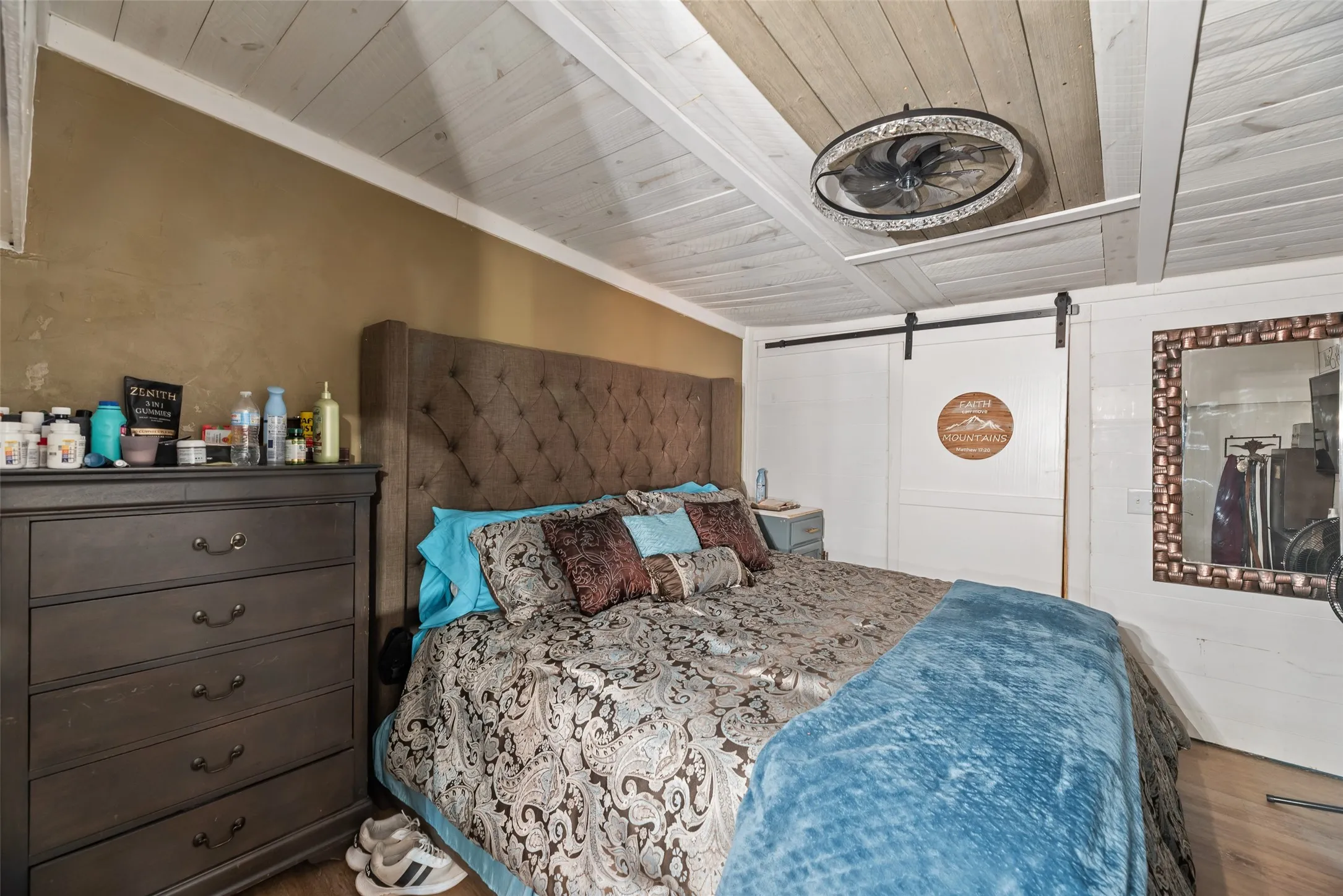 Wood-finish flooring, tufted upholstered headboard, white plank ceiling, and a ceiling fan