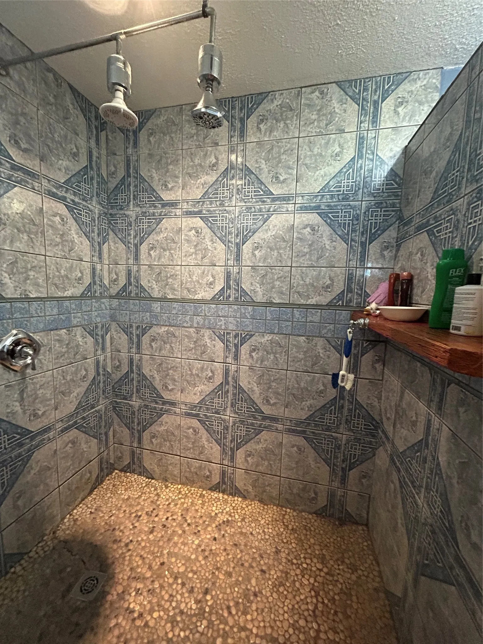 Shower stall featuring patterned ceramic tile walls, a pebble floor, a built-in wood-finish shelf, and dual shower heads