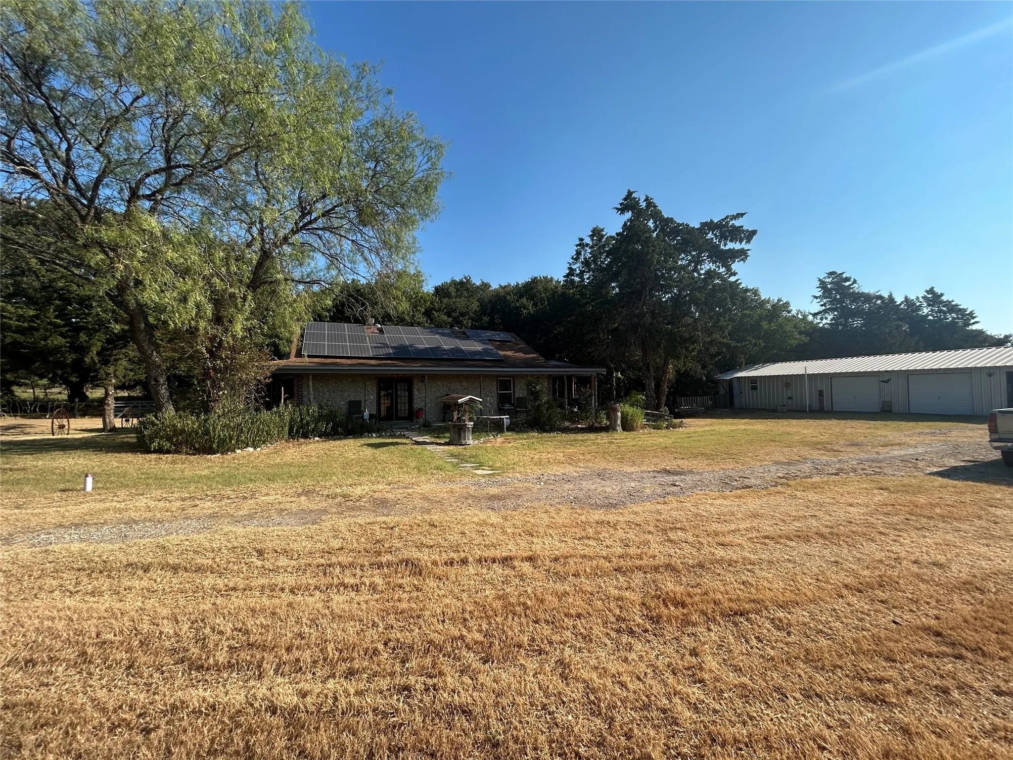 Stone exterior residence with solar panels, covered porch, mature trees, and a detached outbuilding with a metal roof