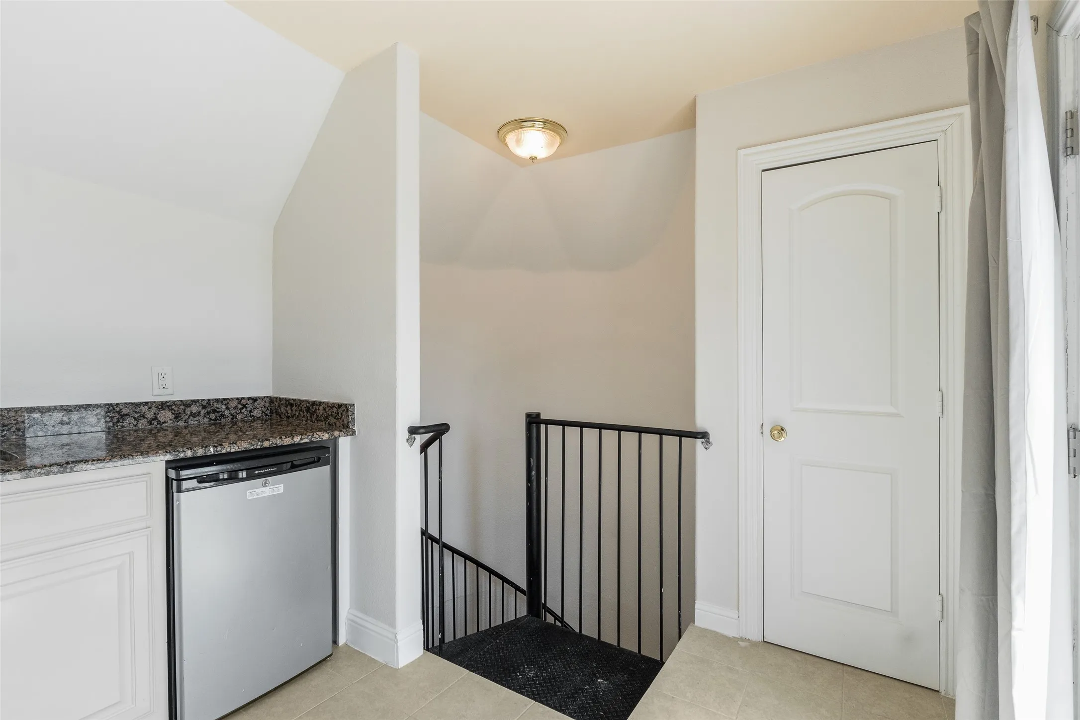 Built-in wet bar with dark granite countertop, under-counter refrigerator, white cabinetry, and adjacent staircase with black metal railing