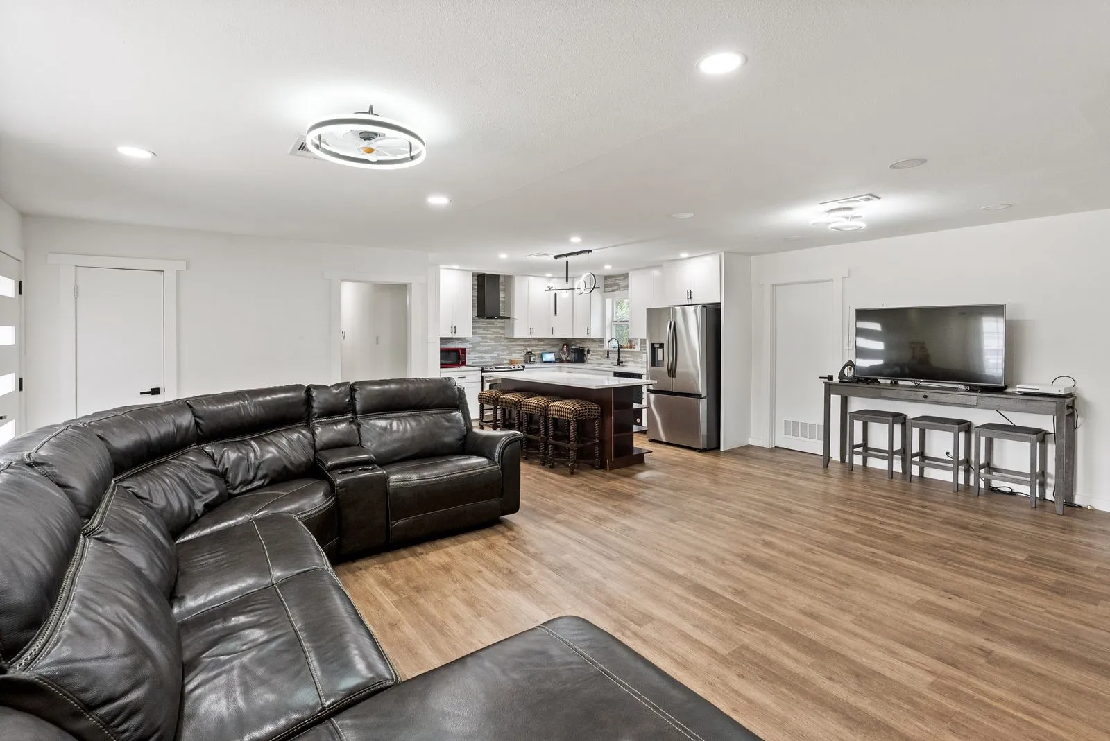 Open-concept living area featuring a curved sectional layout, wood-finish flooring throughout, recessed and pendant lighting, stainless steel appliances, and a kitchen island with seating