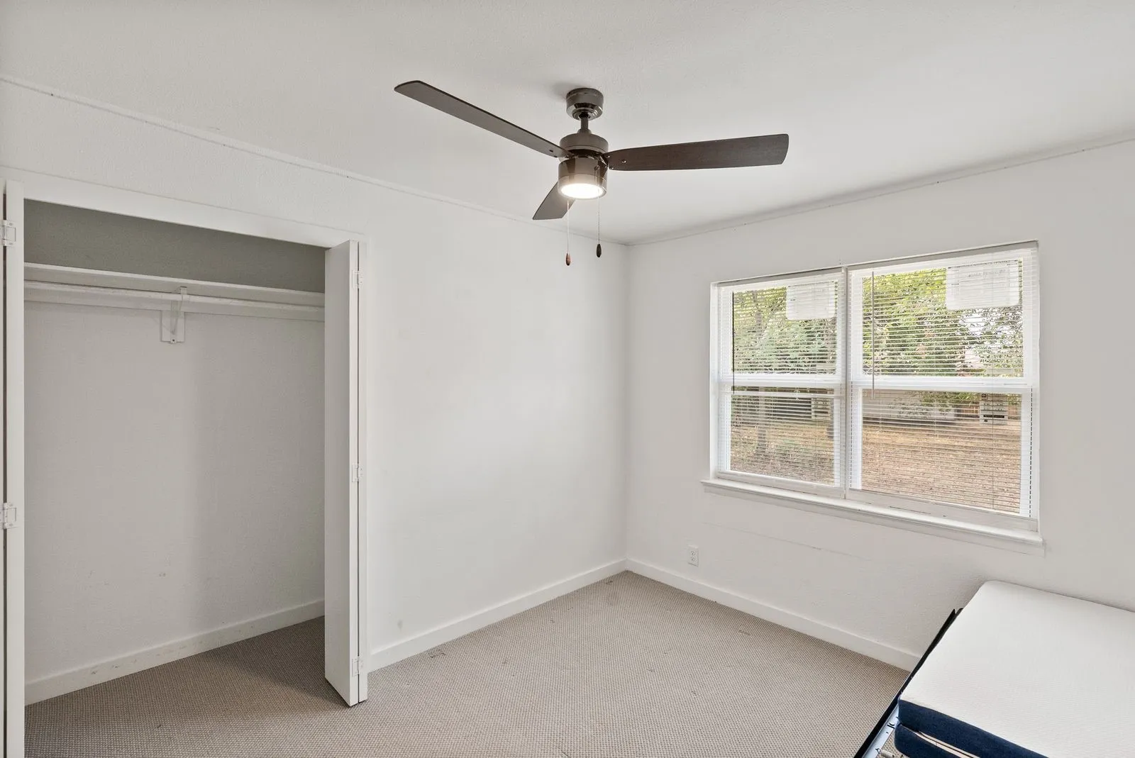 White walls, carpet flooring, window with blinds, ceiling fan with light, and built-in closet with hanging rod