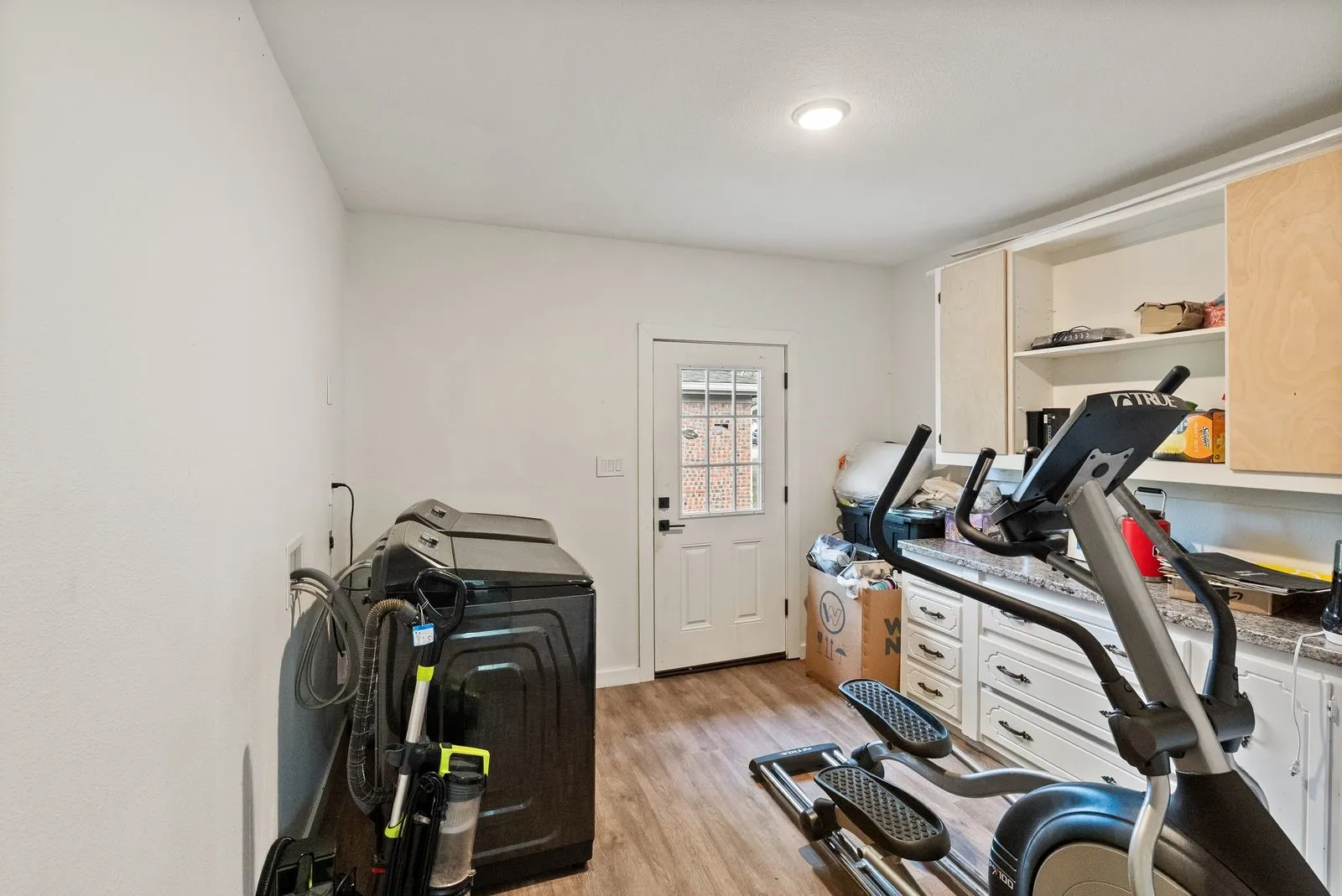 Laundry area with wood-finish flooring, white cabinetry, integrated shelving, and a patterned glass-paneled door