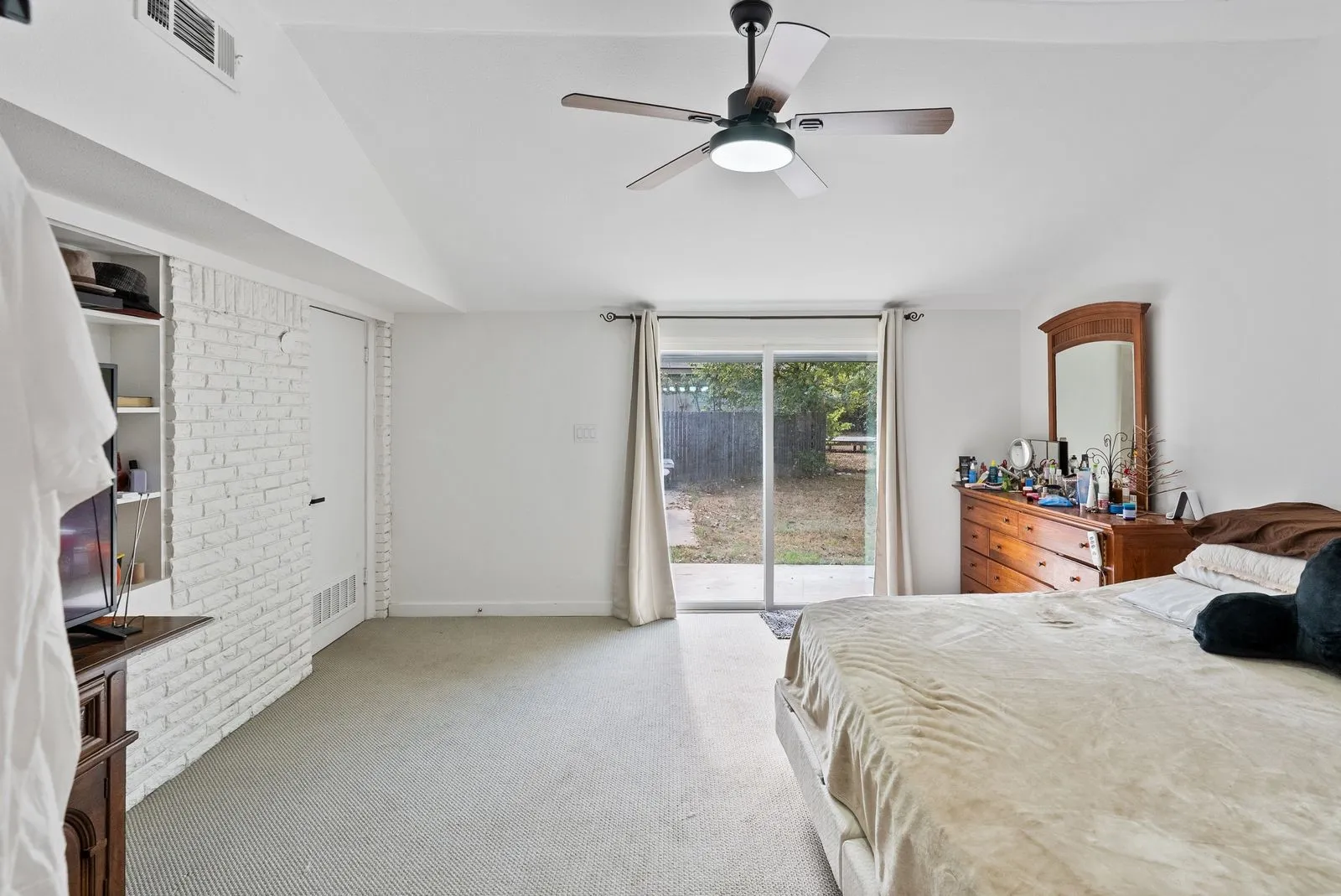 Vaulted ceiling featuring a ceiling fan with integrated lighting, white painted brick accent wall, built-in shelving, and sliding glass doors providing access to the exterior