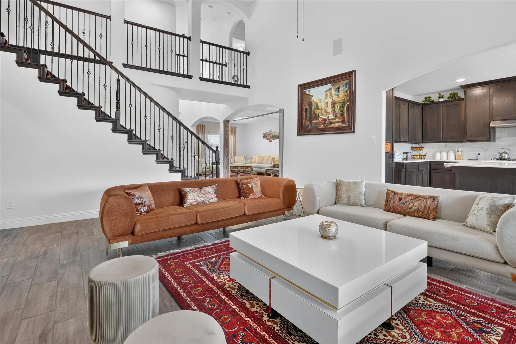 Two-story ceiling, wood-finish flooring, dark wood staircase with wrought iron balusters, arched doorway, and built-in kitchen cabinetry with light-colored backsplash