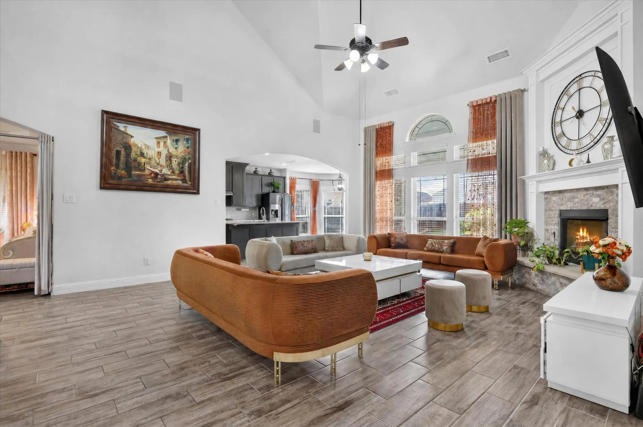 Soaring vaulted ceiling with a ceiling fan, wood-finish flooring, a stone fireplace with a white mantel, and large windows with an arched transom