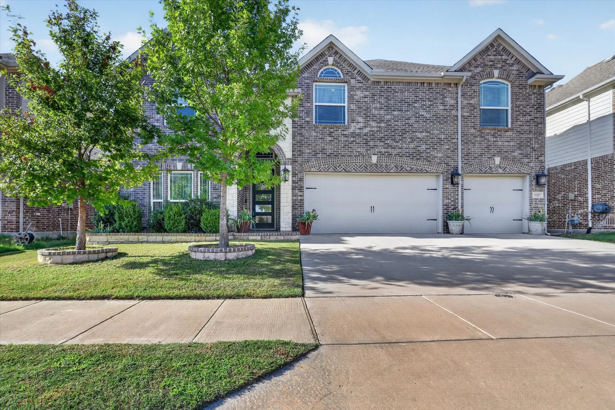 Brick facade with arched entryway, two-car garage, concrete driveway, and landscaped front yard with established trees
