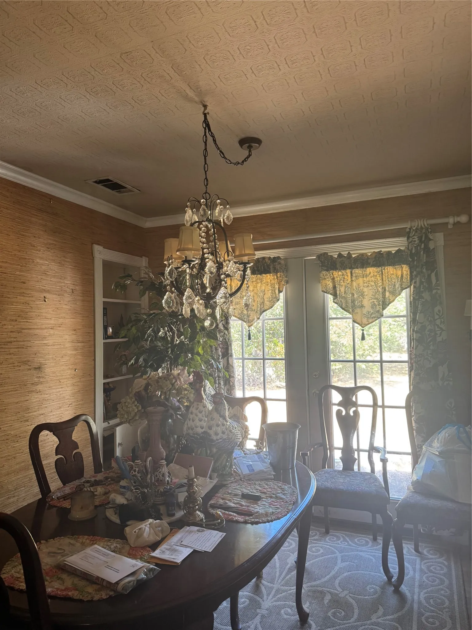 Ceiling-mounted chandelier with crystal accents, crown molding, double French doors, patterned wallpaper, and built-in shelving