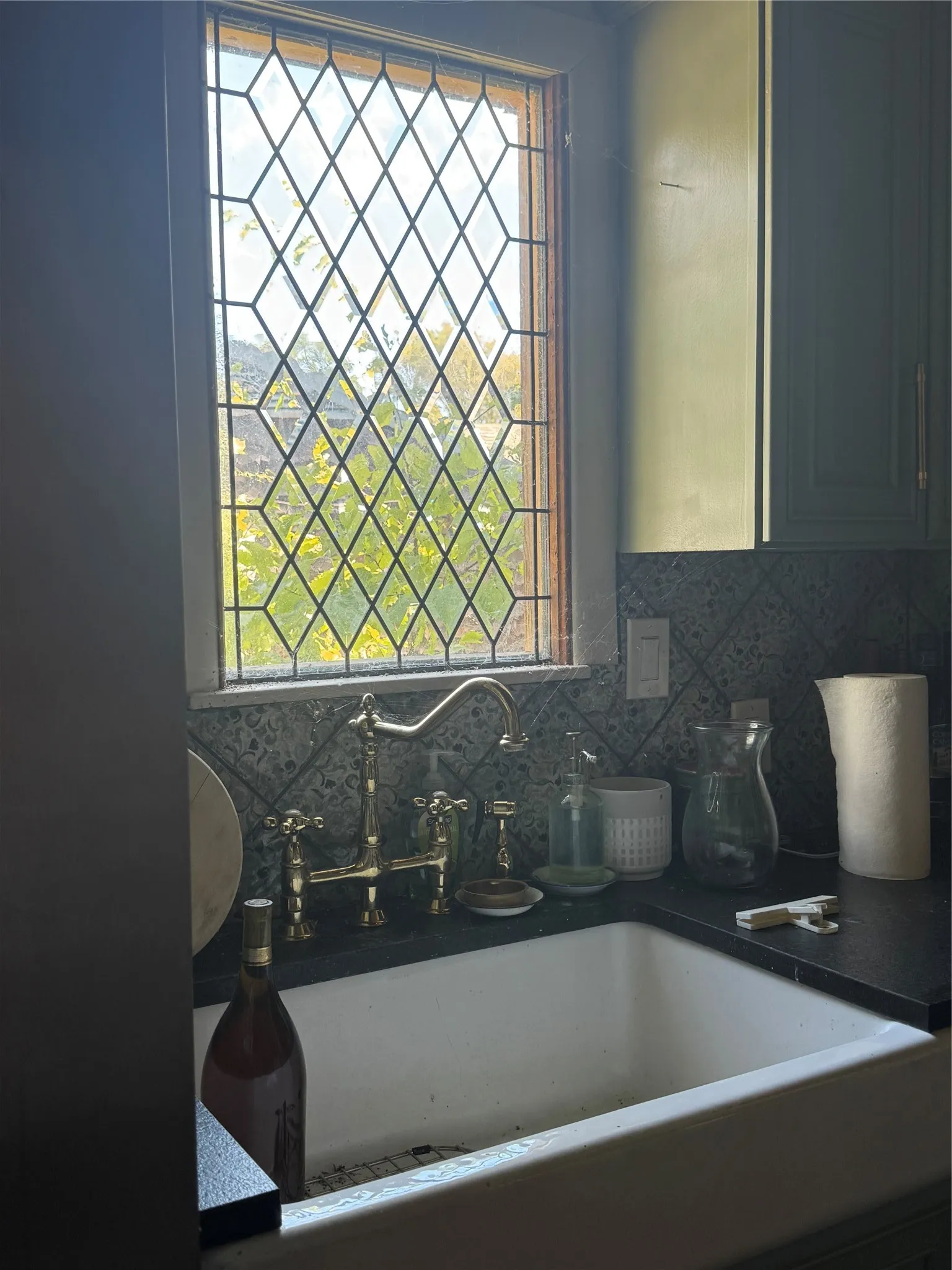 Farmhouse sink with a brass-finish bridge faucet, dark countertop, patterned tile backsplash, and diamond-pane window