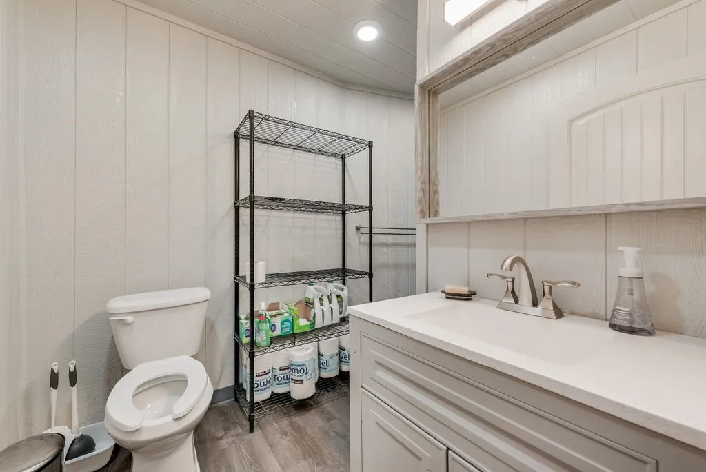 White paneling, wood-finish flooring, light gray vanity with integrated sink, chrome-finish faucet, and overhead vanity lighting
