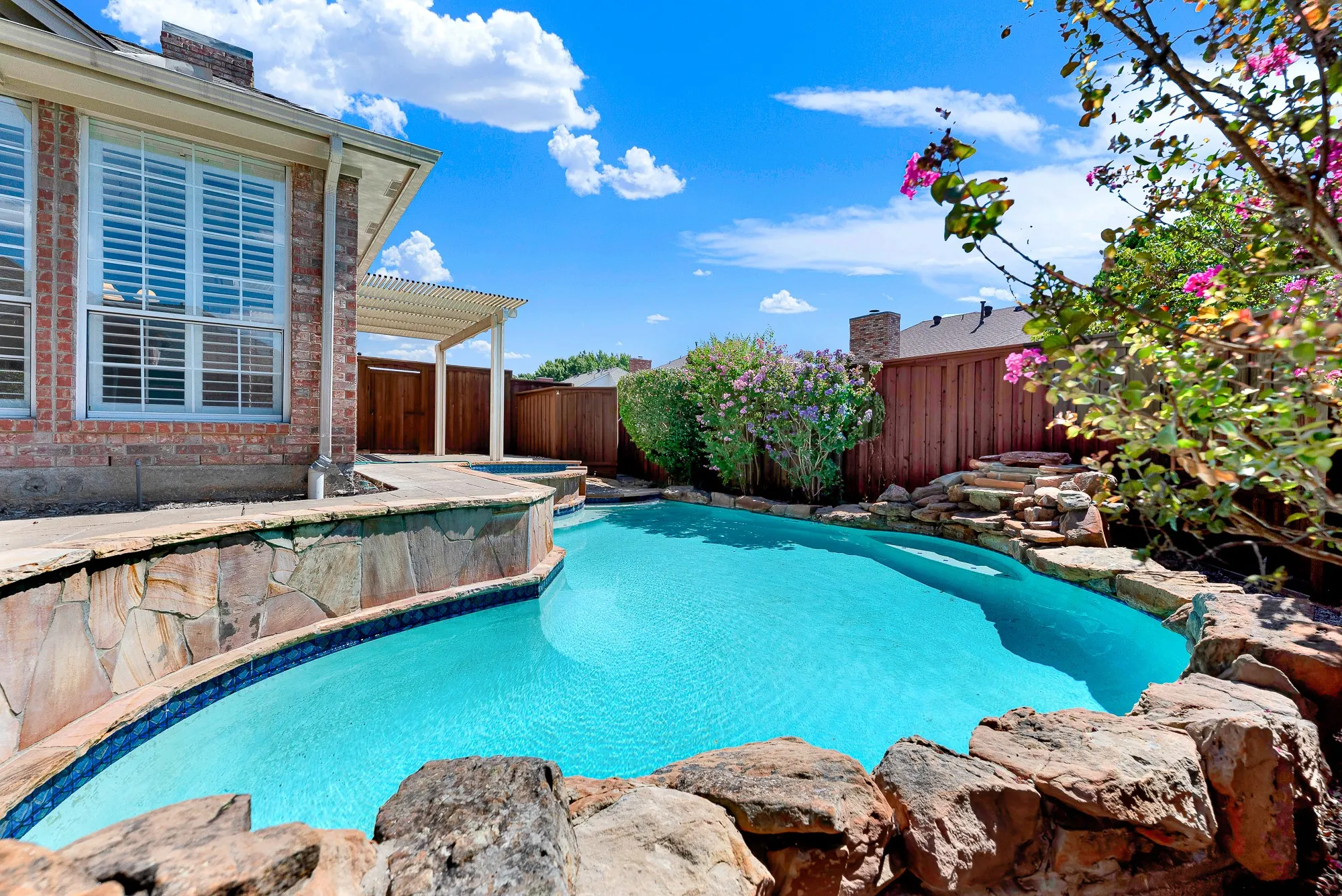 Natural stone rockwork and a gentle waterfall accent the edge of this private backyard pool oasis. Listen to the soothing sound of trickling water while basking in the sun