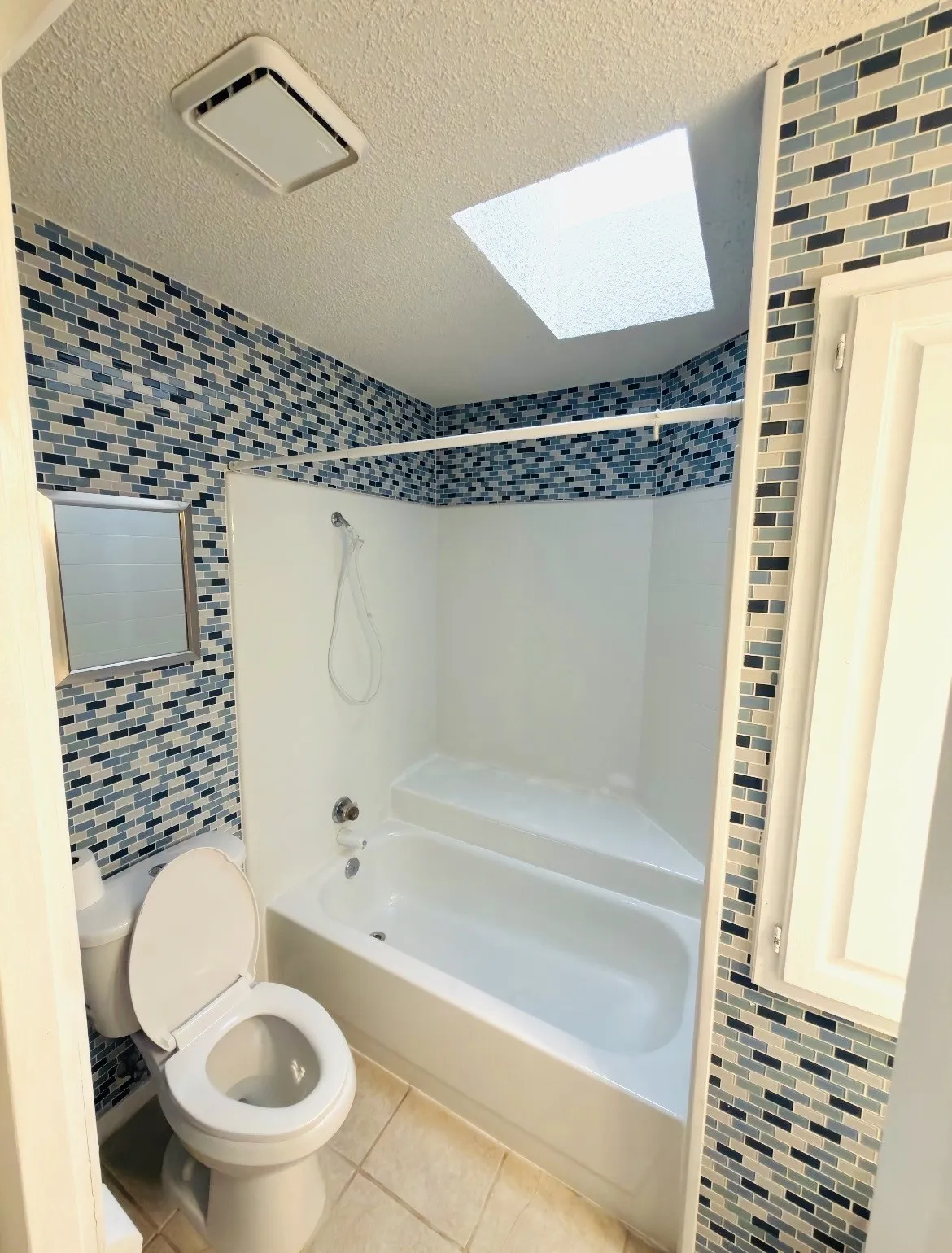 Recessed ceiling light, white bathtub with integrated seating, mosaic tile wall accents, and an overhead ventilation fan