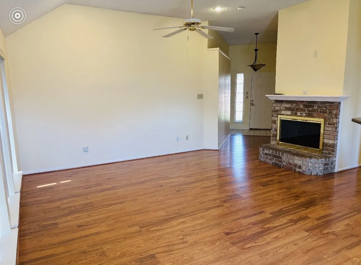 Vaulted ceiling with a ceiling fan, brick fireplace with a mantel, wood-finish flooring, and a decorative pendant light fixture