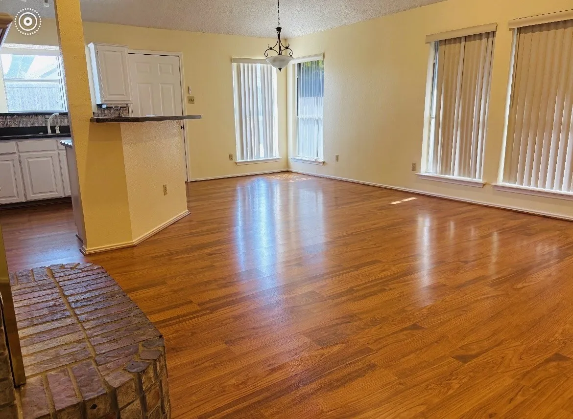 Wood-finish flooring throughout, multiple windows with vertical blinds, brick fireplace hearth, and a built-in kitchen pass-through counter with a dark-toned surface