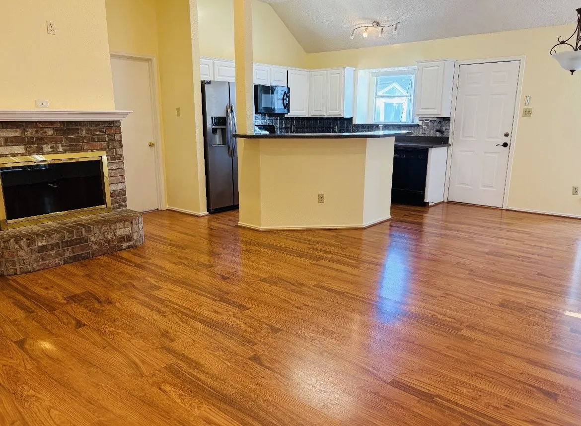 Wood-finish flooring, brick-faced fireplace, vaulted ceiling, white cabinetry, and stainless steel refrigerator